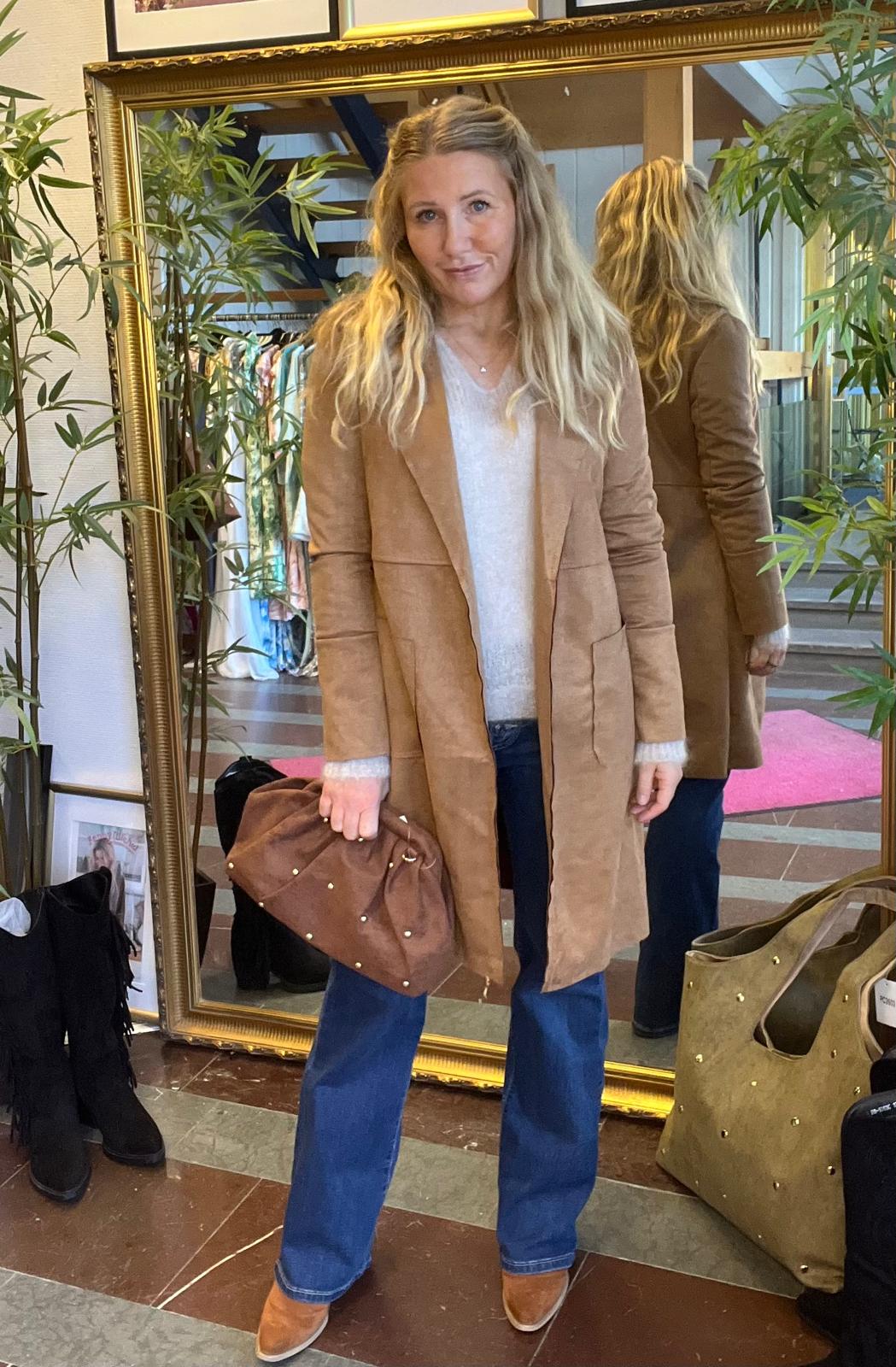 Kvinna med långt blont hår poserar och håller en brun clutch med nitar, klädd i camel kappa, ljus tröja och blå jeans. Hon står framför en stor guldramsspegel i en butiksmiljö med klädställning och växter.