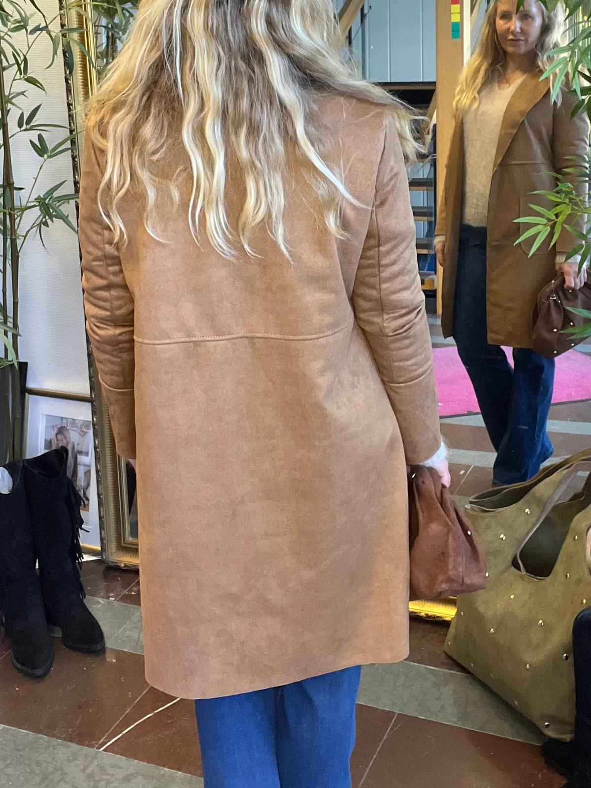 Kvinna med långt blont hår provar en lång beige kappa och står med ryggen mot kameran framför en stor spegel. I spegeln syns hennes ansikte, jeans och en brun handväska i en butiksmiljö.