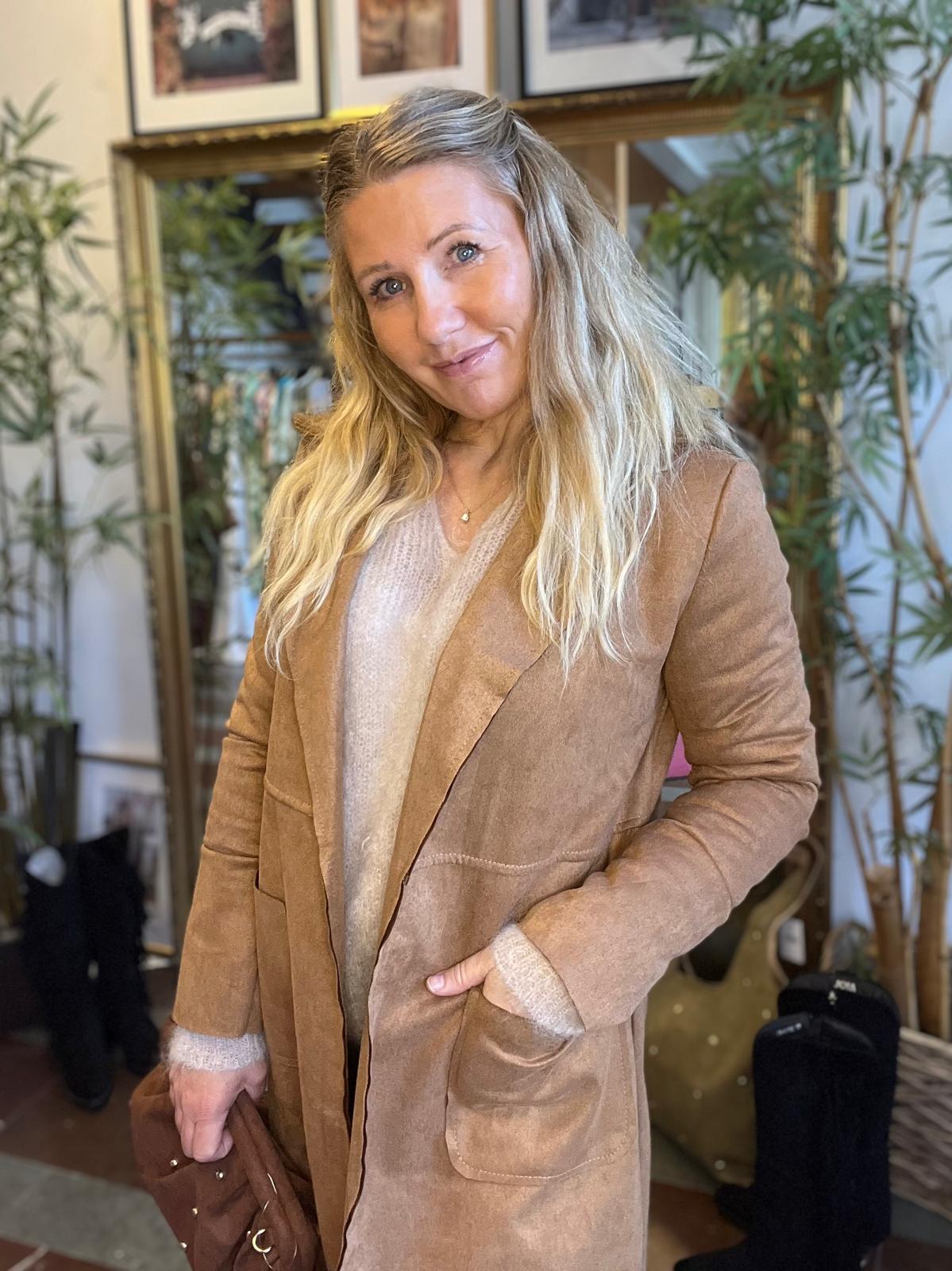 Leende kvinna med långt blont hår poserar inomhus med ena handen i fickan, klädd i ljus stickad tröja och beige mockaliknande kappa. I bakgrunden syns växter, en spegel och inramade bilder.
