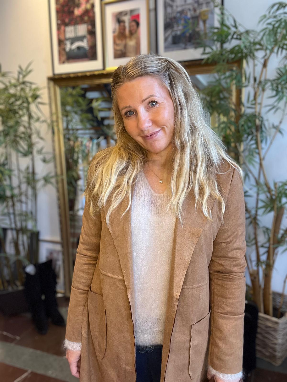 Kvinna med långt blont hår ler mot kameran och står inomhus, klädd i brun kappa och ljus tröja. I bakgrunden syns krukväxter och en hylla med inramade fotografier.