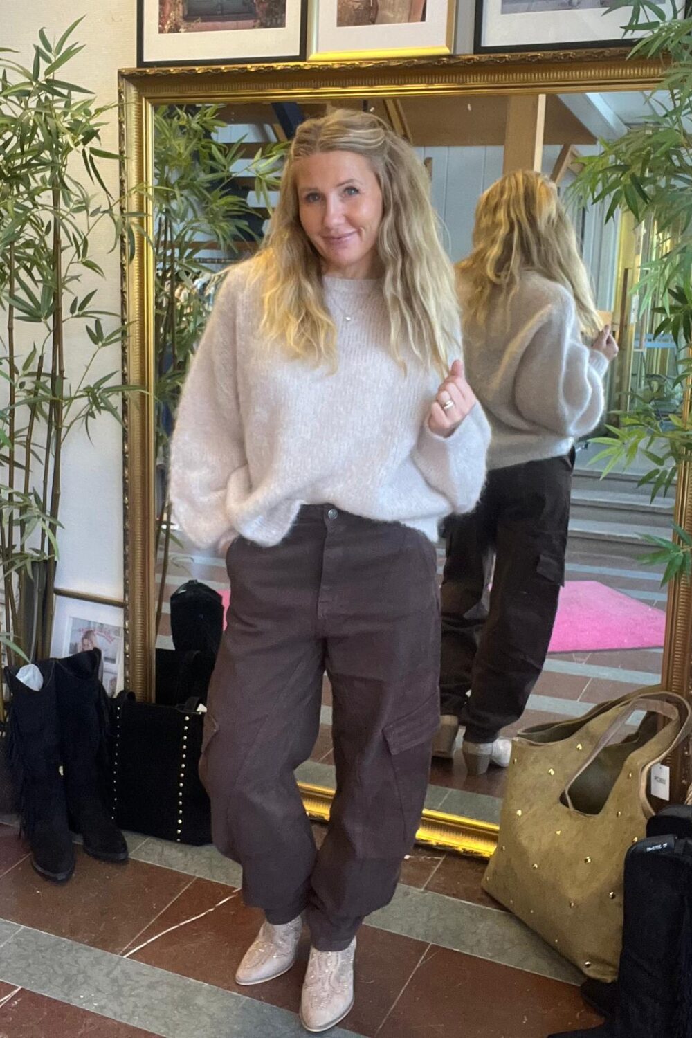 Kvinna med långt blont hår poserar framför en stor guldramsspegel, med ena handen i fickan och den andra lyft. Hon bär ljus stickad tröja, mörka cargobyxor och ljusa boots, omgiven av växter, väskor och stövlar.