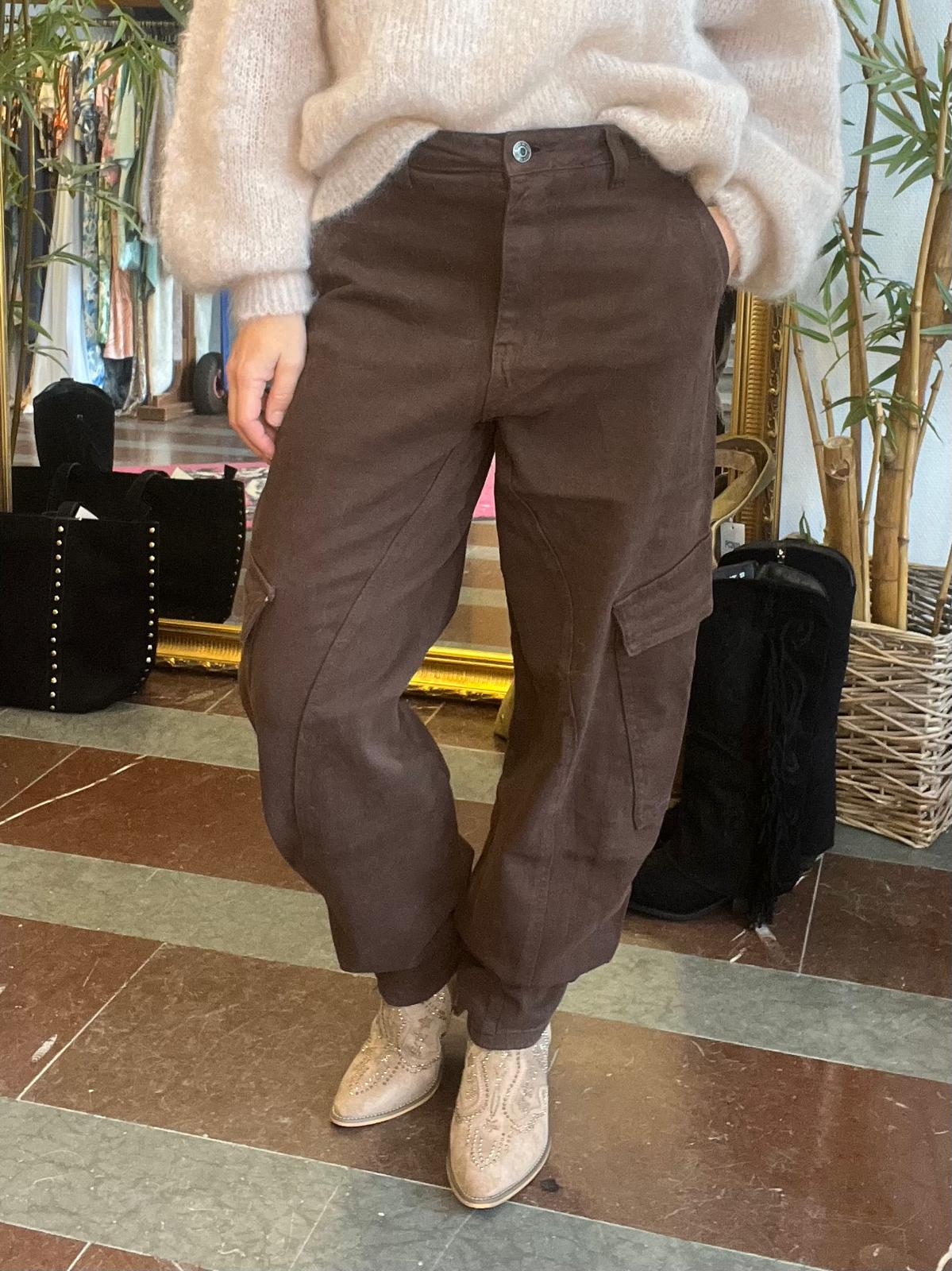 Person står med benen korsade och visar bruna cargobyxor med sidfickor, tillsammans med ljus stickad tröja och beige brogue-kängor. Inomhusmiljö med stor spegel, bambuväxt och svarta väskor i bakgrunden.