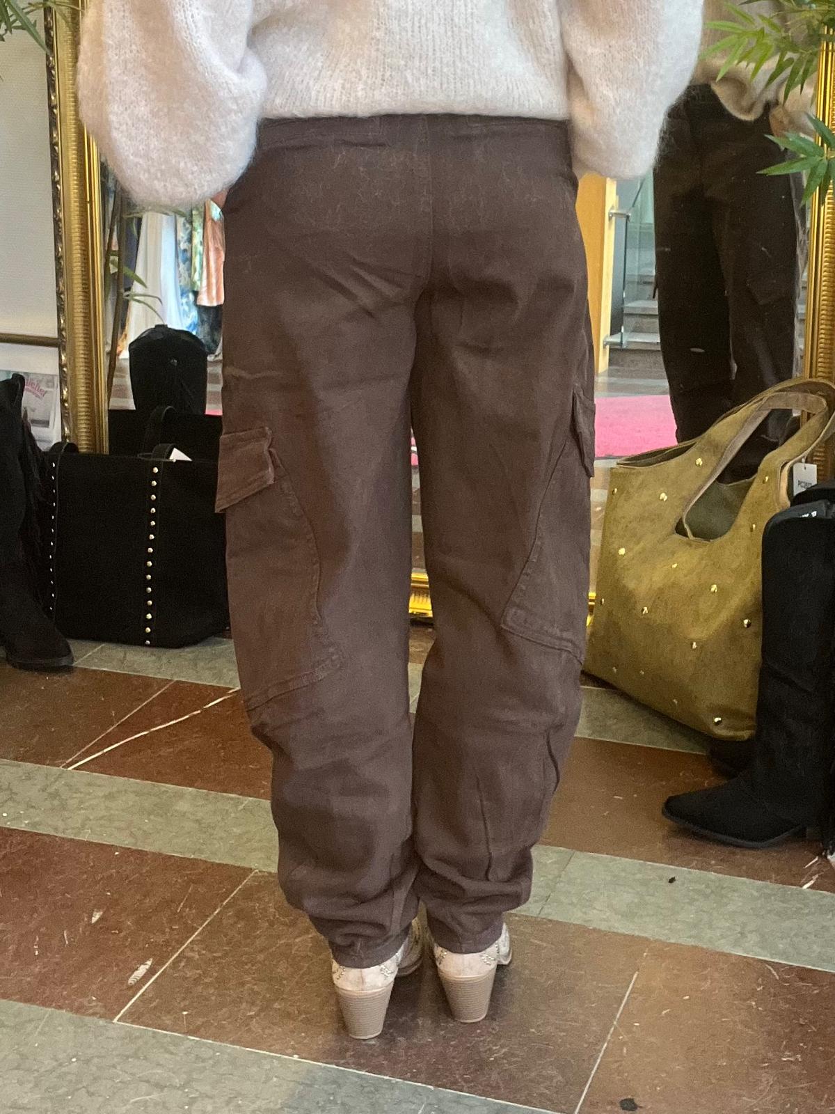 Person står med ryggen mot kameran och provar bruna cargobyxor med sidofickor och vita skor. Hen står framför en stor guldramsspegel i en butik, med väskor och skor synliga på golvet.