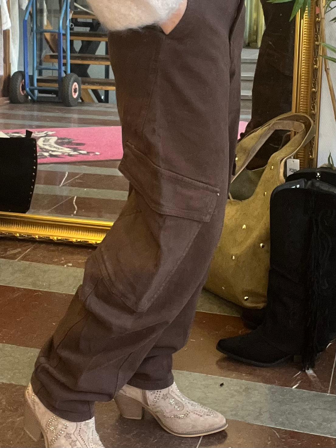 Person står i profil och provar mörkbruna cargobyxor med stora sidofickor, kombinerat med ljusa boots med dekor. Miljön liknar en butik eller showroom med spegel, väska och möbler i bakgrunden.