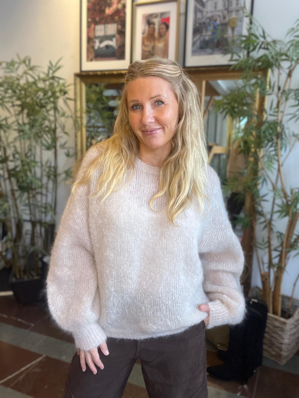 Leende kvinna med långt blont hår poserar med ena handen i fickan, klädd i ljus stickad tröja och mörka byxor. Hon står inomhus framför stora gröna växter och en hylla med inramade foton.