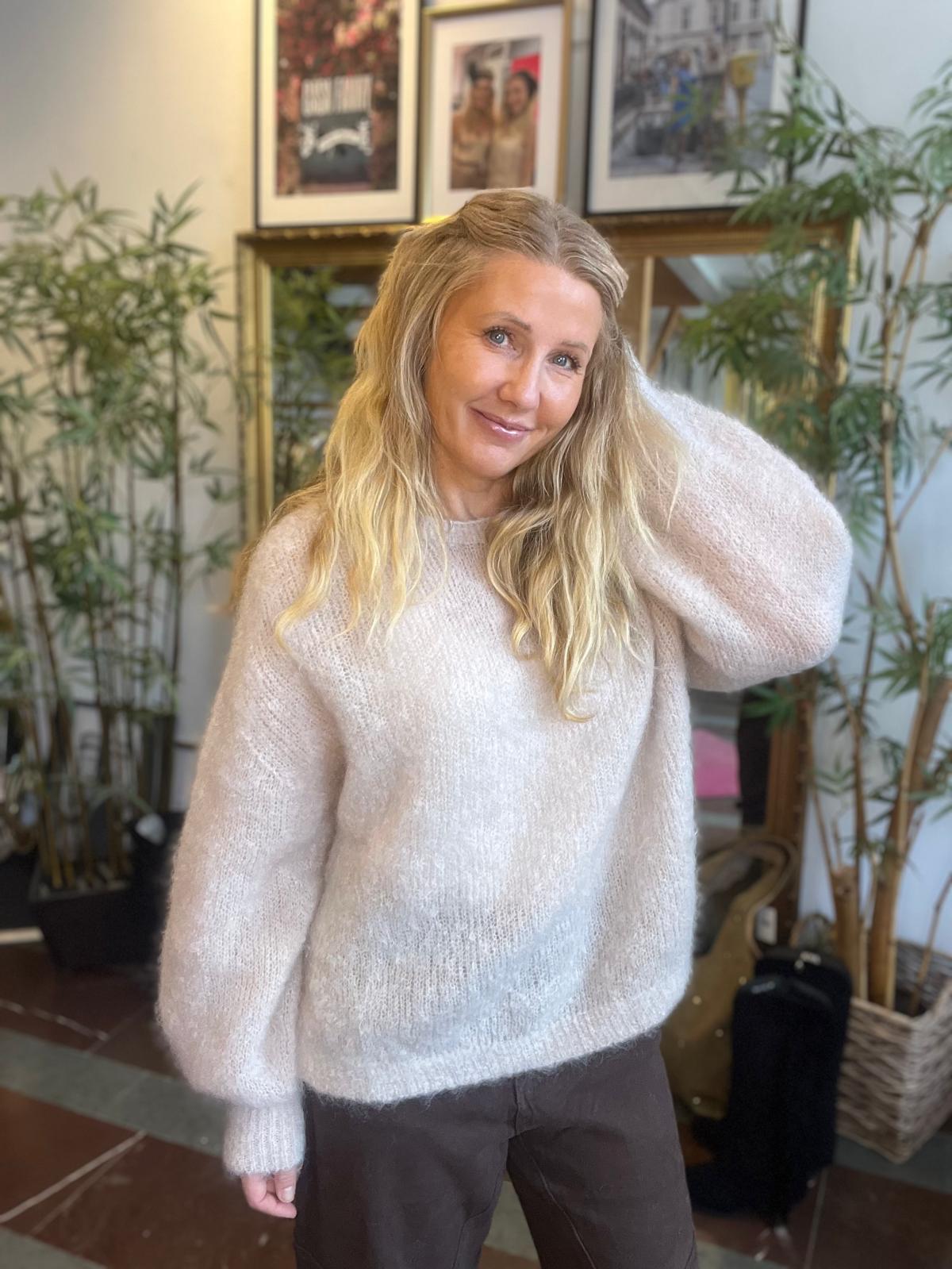 Leende kvinna med långt blont hår står och håller ena handen vid håret, klädd i ljus stickad tröja och mörka byxor. Inomhusmiljö med gröna växter och inramade foton på väggen bakom.