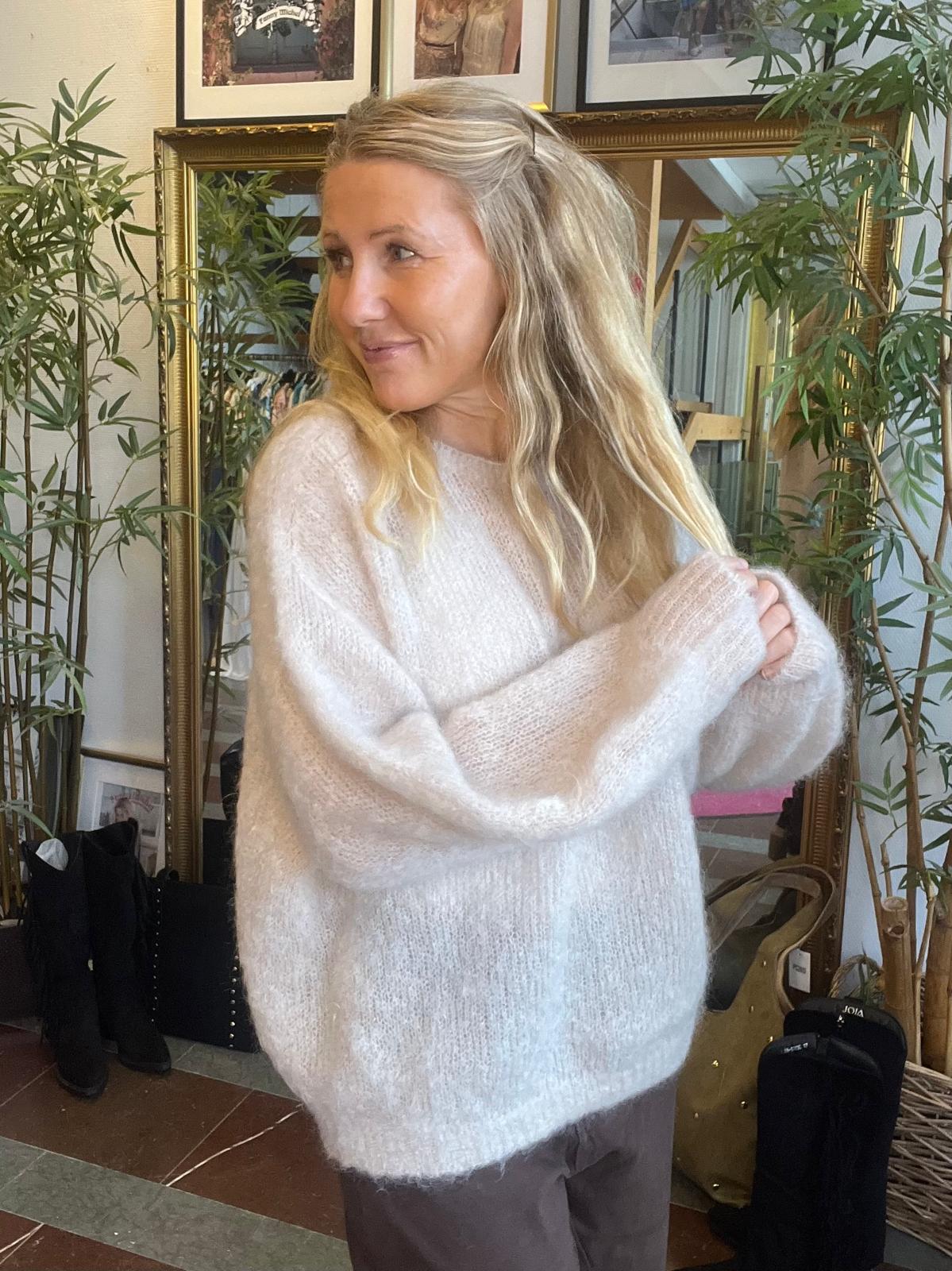 Blond kvinna står inomhus och drar handen genom håret medan hon ser åt sidan med ett litet leende. Hon bär en ljus, fluffig stickad tröja; bakom syns spegel, gröna växter och skor.