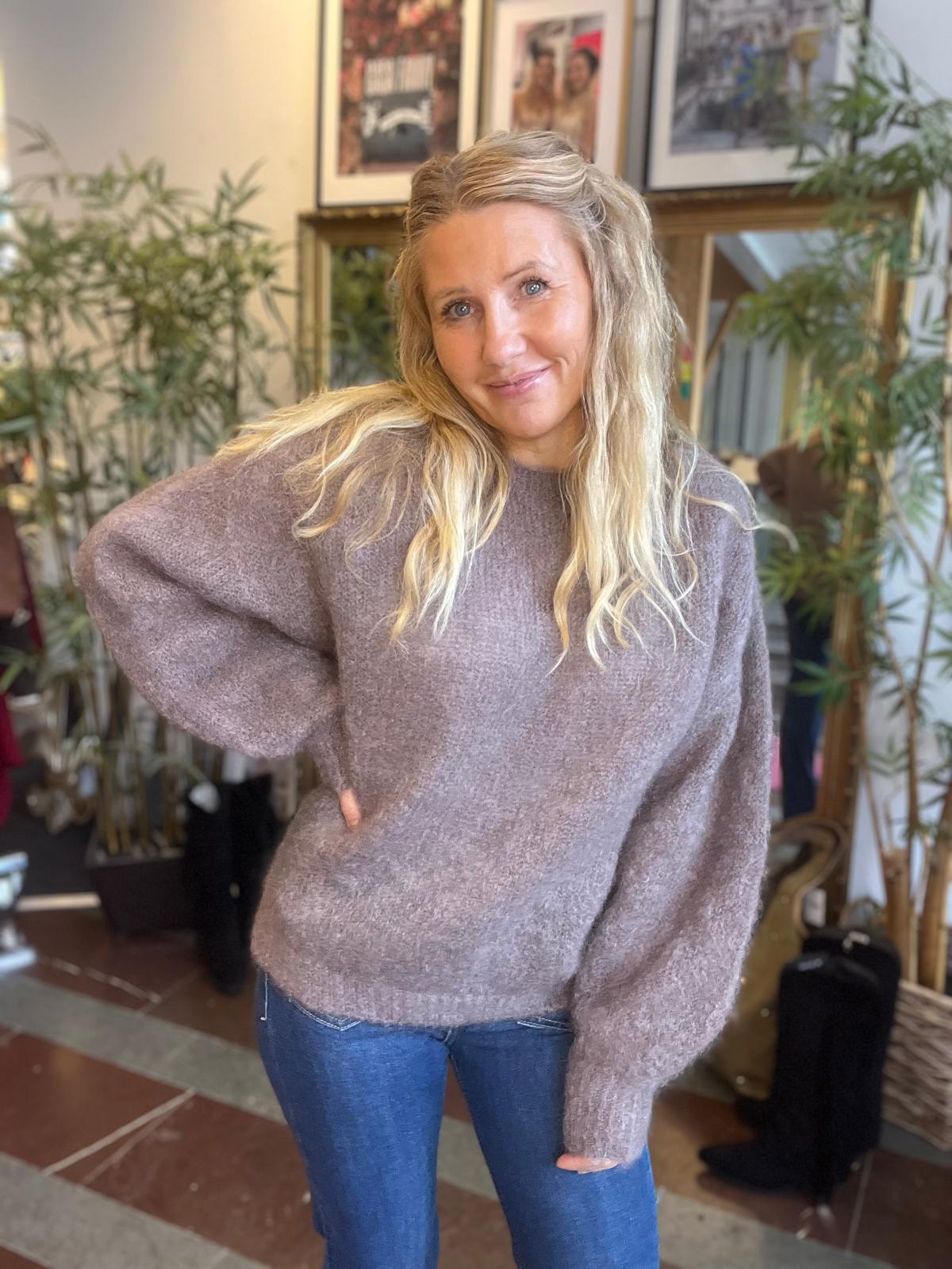 Kvinna med långt blont hår står och ler mot kameran med handen på höften. Hon bär en lila stickad tröja och blå jeans, i ett rum med gröna växter och inramade tavlor i bakgrunden.