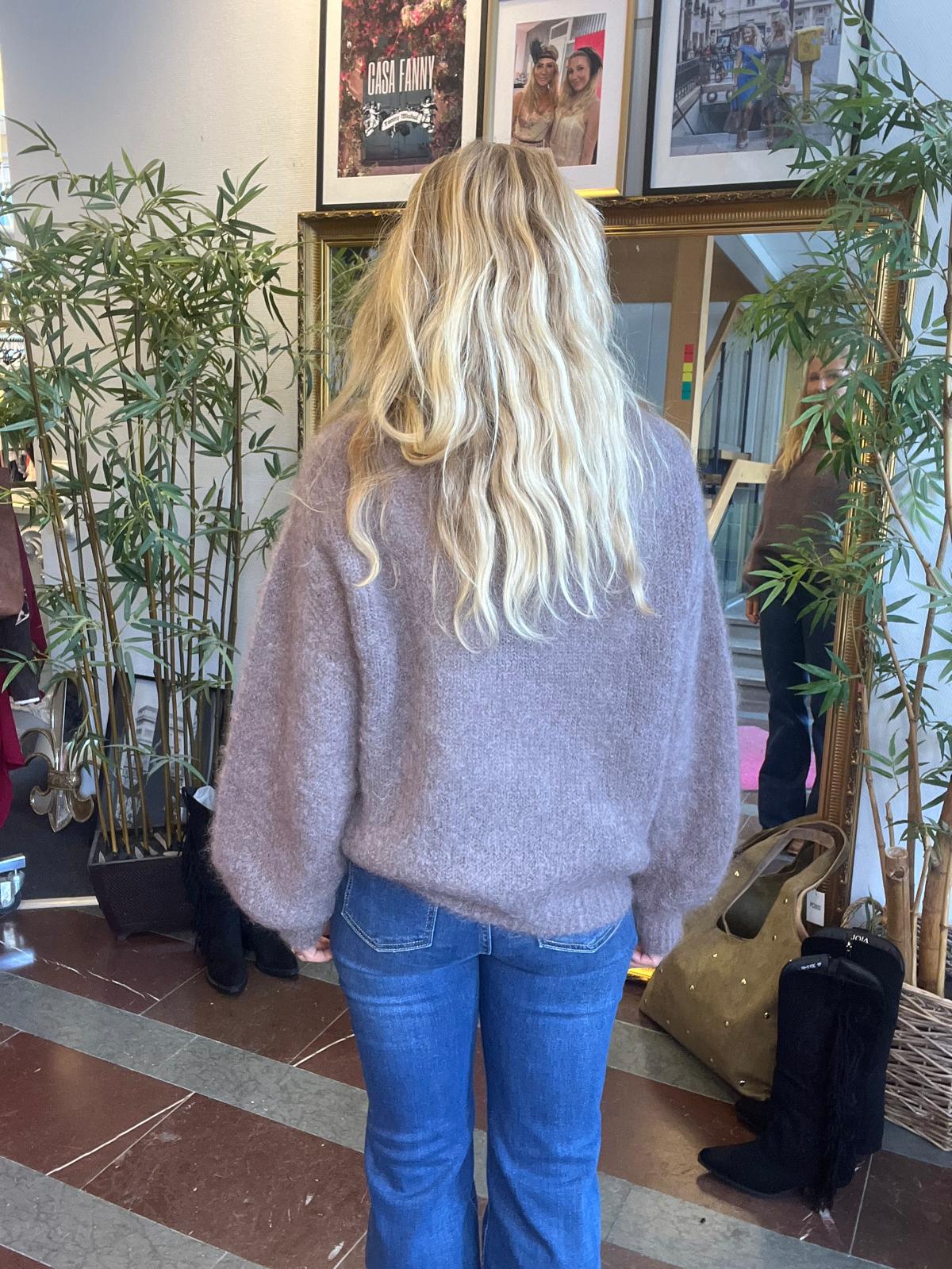 Kvinna med långt blont hår står med ryggen mot kameran och tittar i en stor guldramsspegel i en hall. Bambuväxter och väskor omger spegeln; ovanför hänger tavlor med texten “GASA FANNY”.