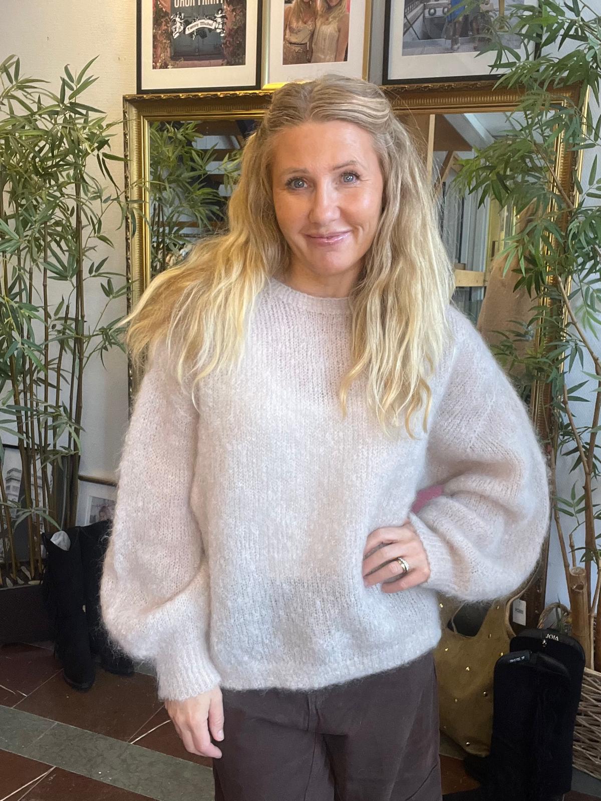 Leende kvinna med långt blont hår står med handen på höften och tittar mot kameran, klädd i ljus, fluffig stickad tröja och mörka byxor. Inomhusmiljö med spegel, bambuväxter och inramade foton bakom.