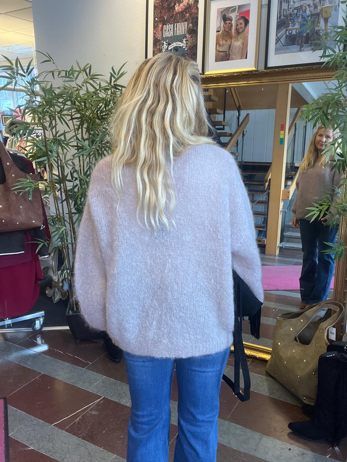 Kvinna med långt blont hår står med ryggen mot kameran och tittar i en stor spegel i en butik. Hon bär lila stickad tröja och jeans; runtom syns kläder, växter och foton samt texten ”CASA FANNY”.