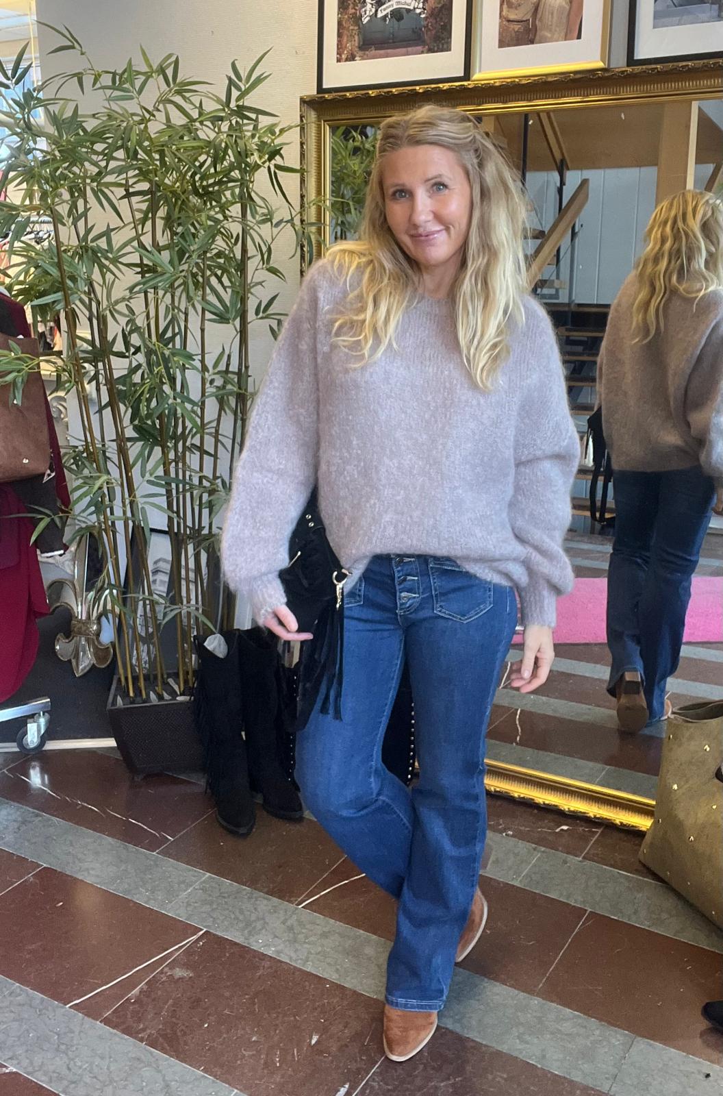 Blond kvinna står och poserar avslappnat med benen i kors, iförd ljusgrå stickad tröja, blå jeans och bruna boots. Inomhusmiljö med stor guldramsspegel som visar hennes rygg, bambuplanta och väskor i bakgrunden.
