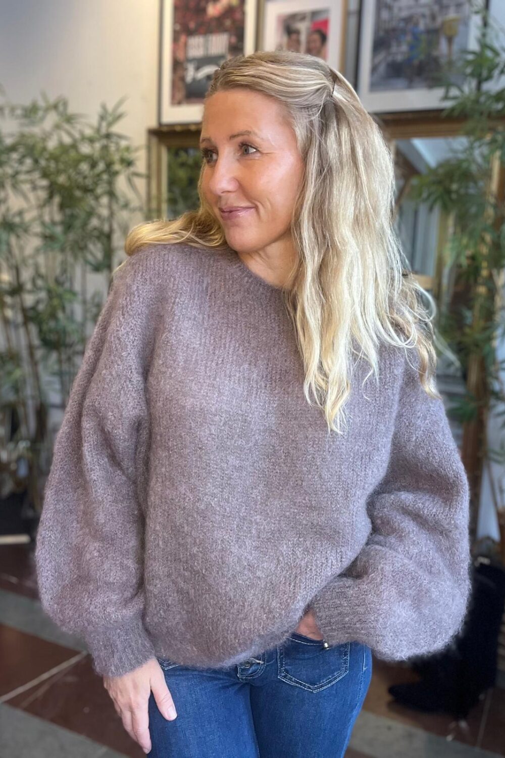 Blond kvinna i lila stickad tröja och blå jeans står inomhus med ena handen i fickan och ser åt sidan med ett lätt leende; bakom henne syns gröna krukväxter och inramade tavlor.