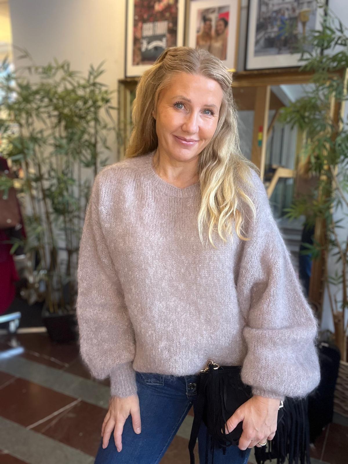 Leende kvinna med långt blont hår står avslappnat och tittar mot kameran, klädd i ljusrosa stickad tröja och blå jeans. Inomhusmiljö med stora gröna växter och inramade bilder på väggen i bakgrunden.