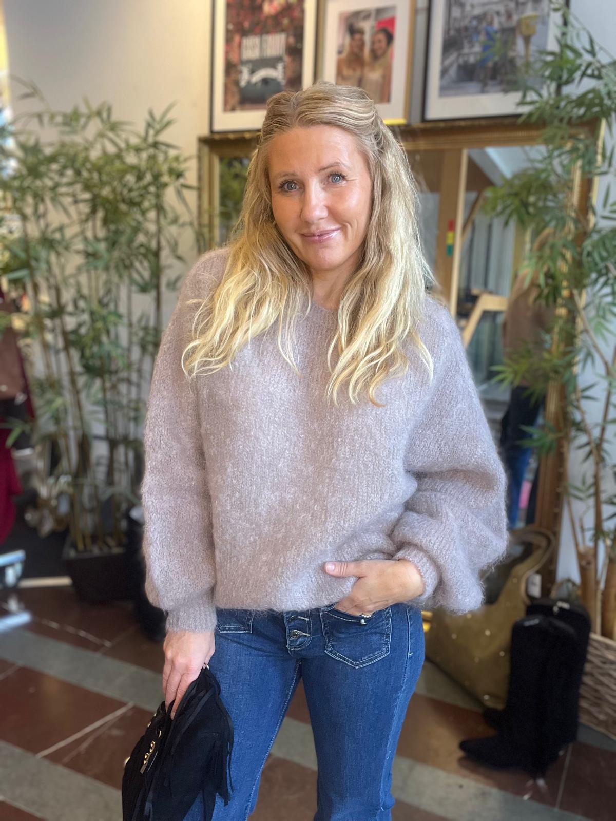 Kvinna med långt blont hår ler mot kameran och står med ena handen i jeansfickan, den andra håller en svart jacka. Hon bär en lila stickad tröja, inomhus bland gröna växter och inramade tavlor.
