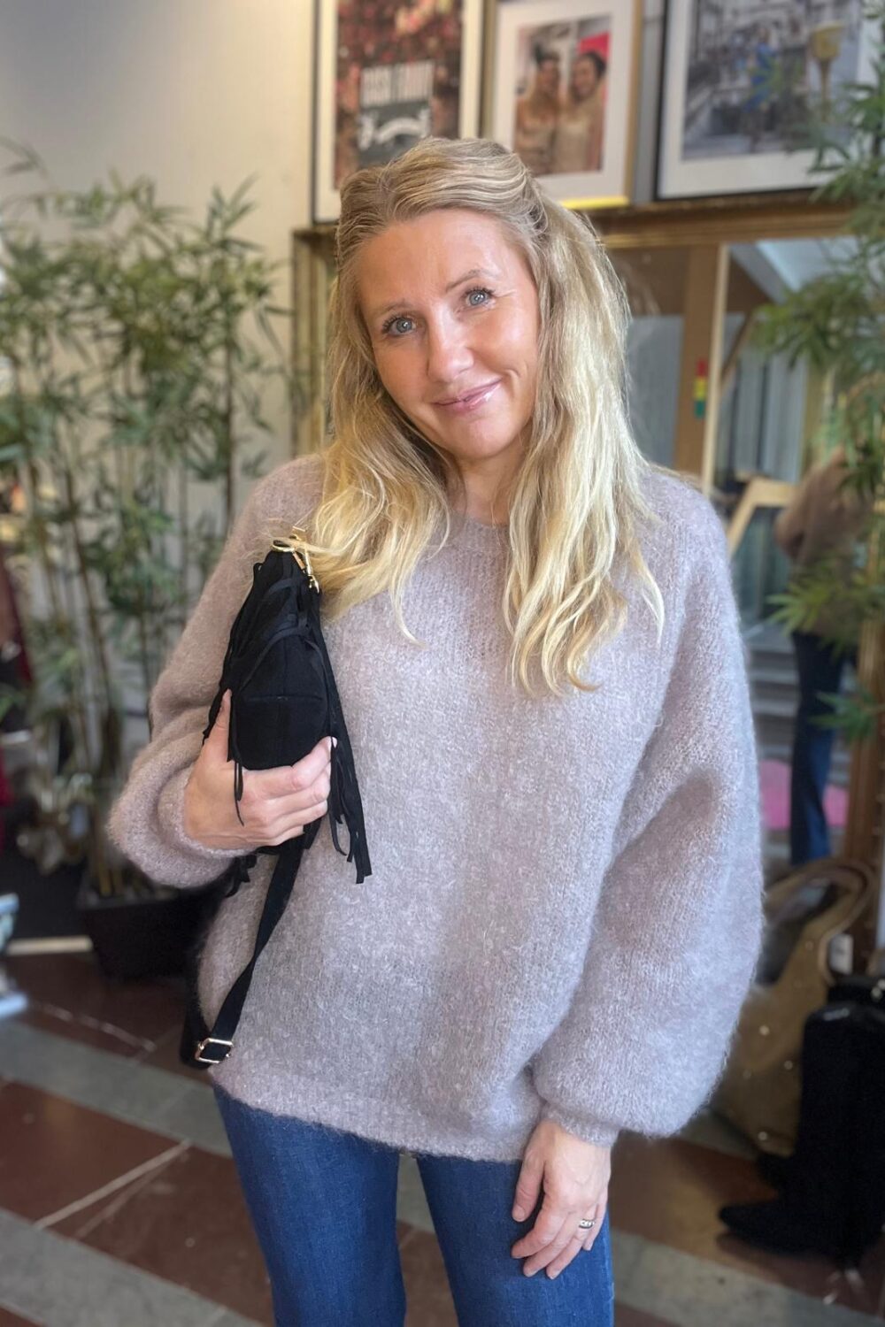Leende kvinna med långt blont hår står och poserar, håller en svart väska mot bröstet. Hon bär en ljus, mjuk stickad tröja och jeans, i en inomhusmiljö med växter och inramade bilder i bakgrunden.