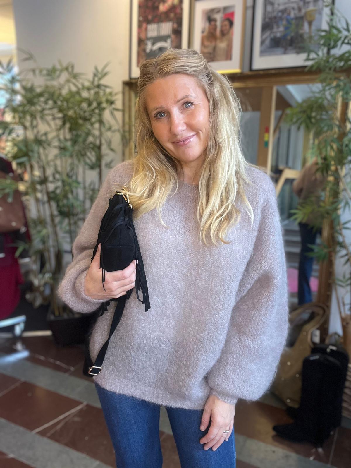Leende kvinna med långt blont hår står och poserar, håller en svart väska mot bröstet. Hon bär en ljus, mjuk stickad tröja och jeans, i en inomhusmiljö med växter och inramade bilder i bakgrunden.