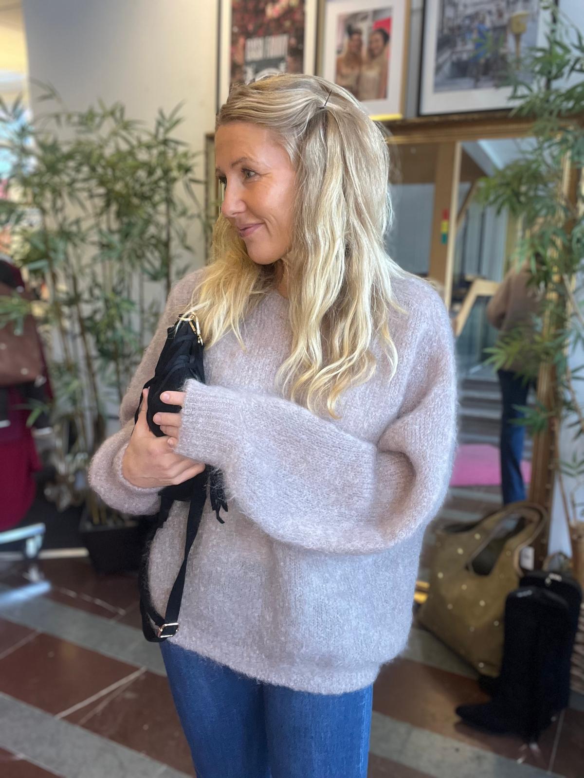 Blond kvinna i ljus stickad tröja står inomhus och håller en svart axelväska mot bröstet medan hon ler och tittar åt sidan. I bakgrunden syns gröna krukväxter, inramade foton och resväskor.