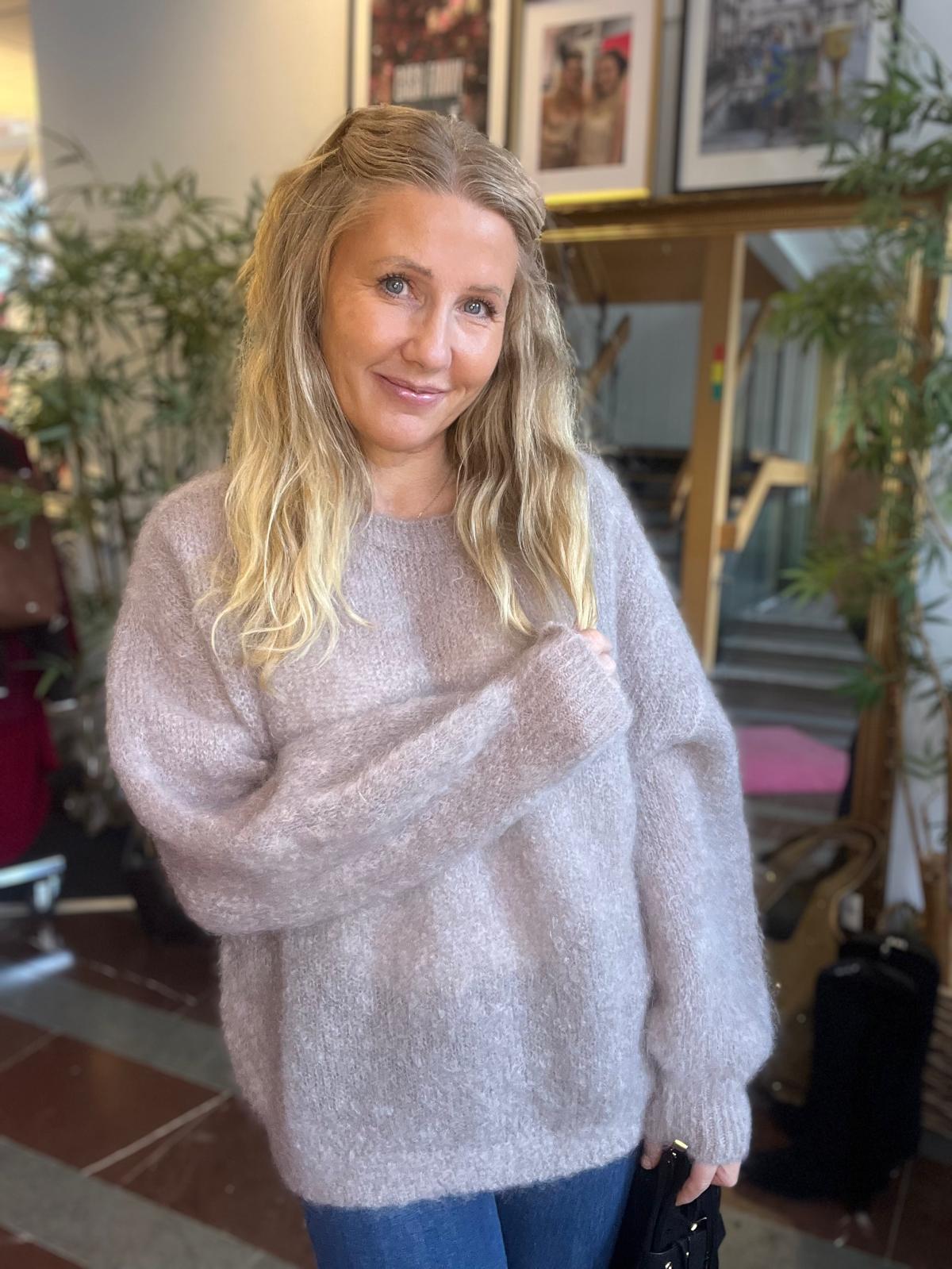 Leende kvinna med långt blont hår står inomhus och ser mot kameran, klädd i ljus, stickad tröja och jeans. Hon håller i tröjärmen; bakom syns gröna växter och inramade tavlor.