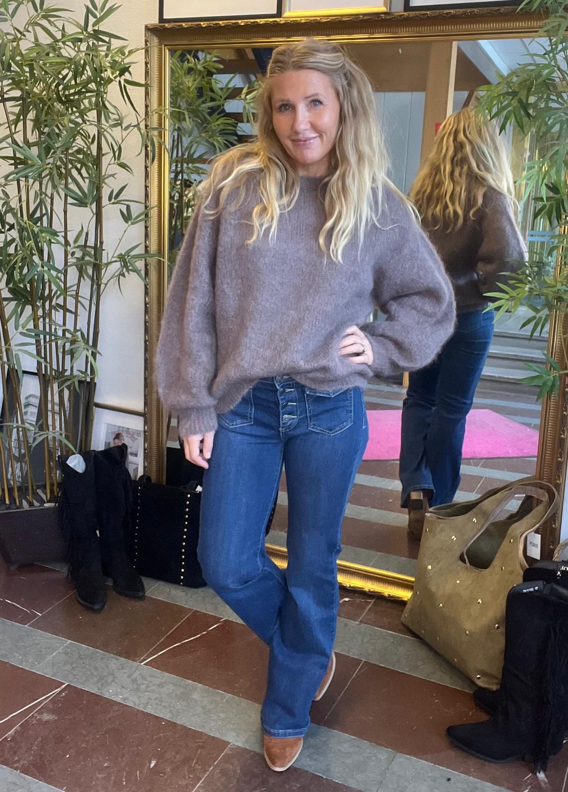 Kvinna med långt blont hår poserar med handen på höften och benet korsat, klädd i grå stickad tröja och blå jeans, framför en stor guldramsspegel. Gröna växter, boots och väskor syns på golvet.