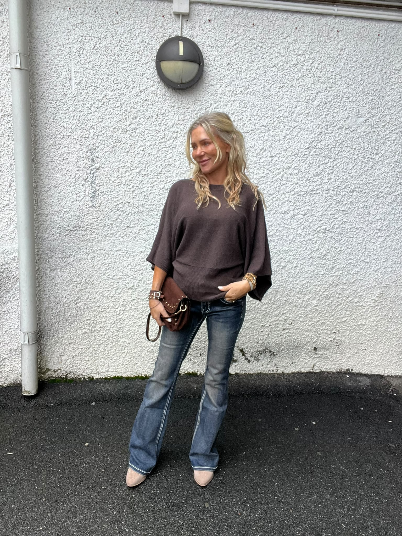 Blond kvinna står lutad mot en vit, grovputsad vägg och tittar åt sidan med ett leende. Hon bär mörk topp, utsvängda jeans och ljusa skor, och håller en brun väska; rund vägglampa ovanför.