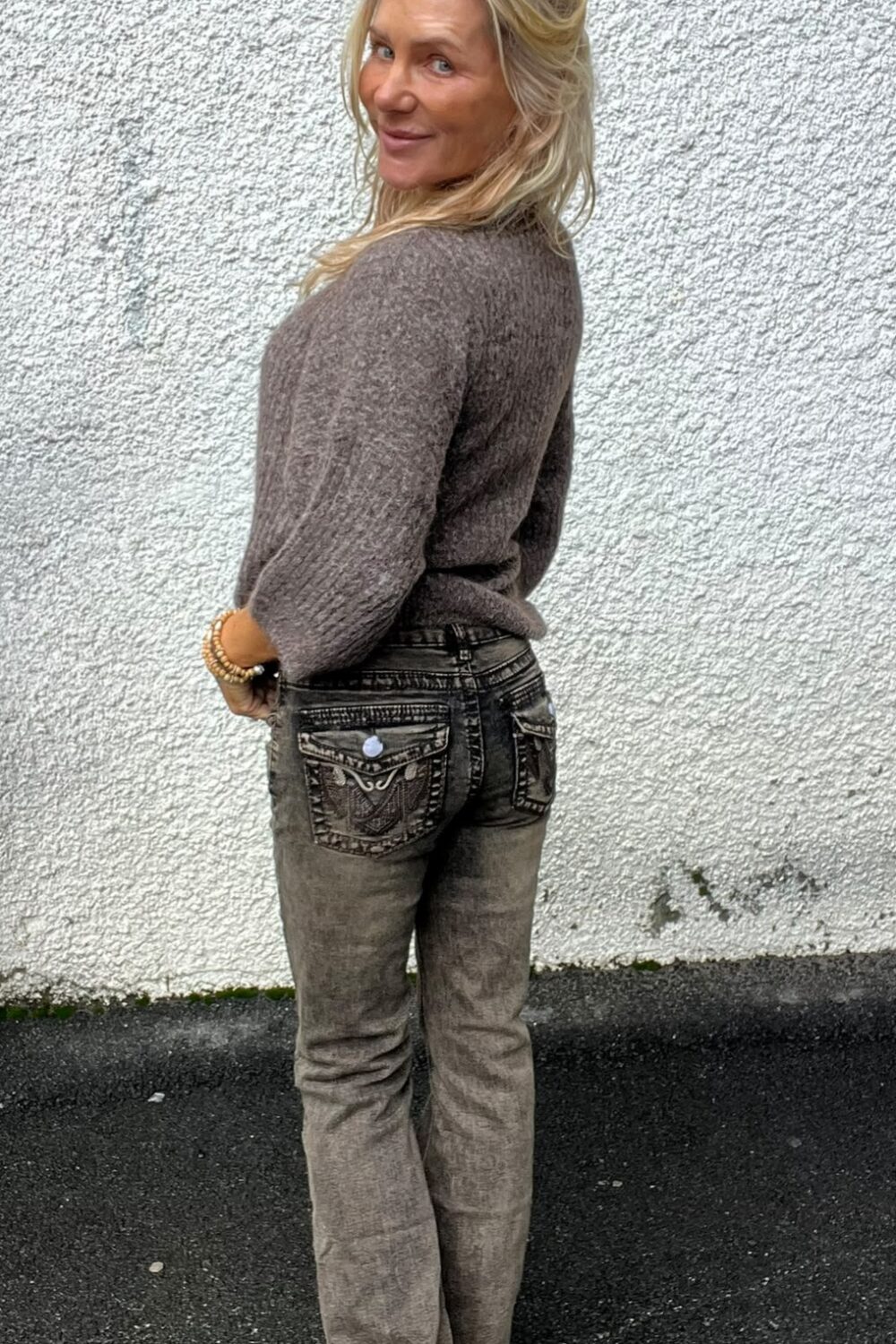 Blond kvinna står utomhus framför en vit, grovputsad vägg och vänder sig om med ett leende mot kameran. Hon bär grå stickad tröja och grå jeans med dekorativa bakfickor, samt armband.