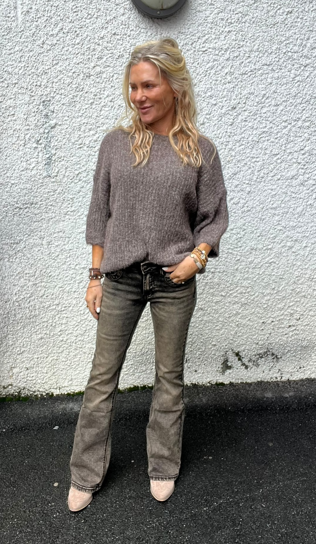 Kvinna med långt blont hår står lutad mot en vit, grovputsad vägg och tittar åt sidan. Hon bär grå stickad tröja, grå jeans och ljusa boots, med handen i fickan och flera armband.