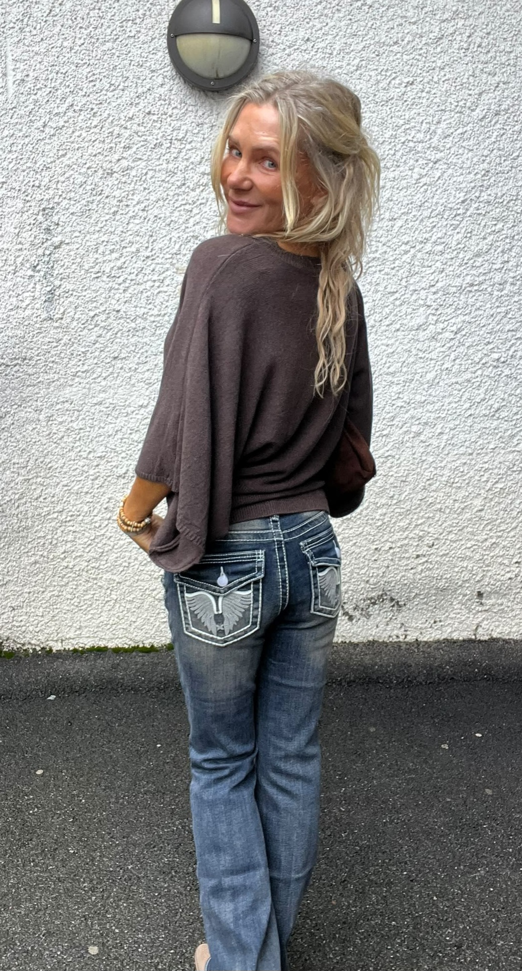 Blond kvinna står med ryggen mot kameran och vänder sig om med ett leende. Hon bär brun stickad tröja och blå jeans med broderade bakfickor, framför en vit putsad vägg med rund utelampa.