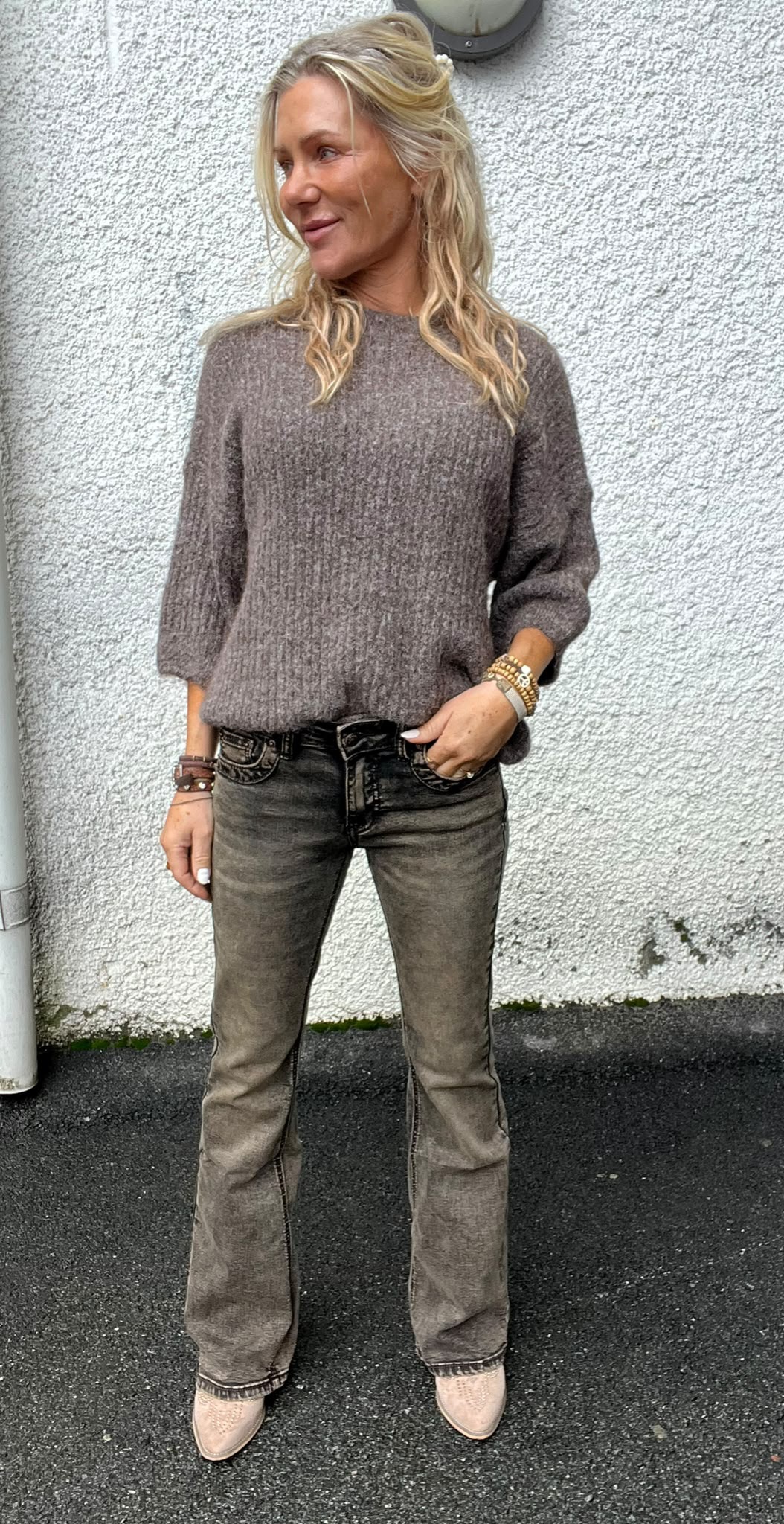 Blond kvinna står avslappnat med ena handen i fickan och tittar åt sidan, klädd i grå stickad tröja, grå jeans och ljusa boots. Hon poserar utomhus mot en vit putsad vägg, med armband på handlederna.