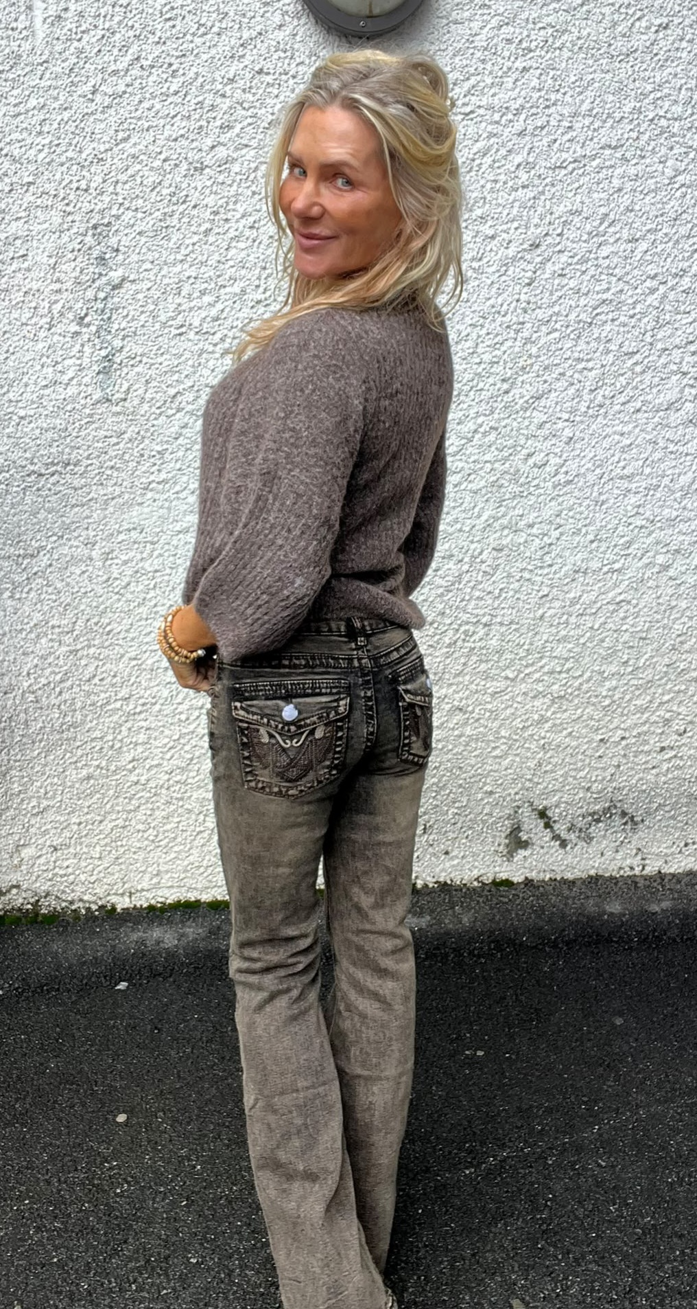 Kvinna med långt blont hår står i profil och vänder sig om mot kameran med ett leende. Hon bär grå stickad tröja och slitna jeans med broderade bakfickor, framför en vitputsad vägg utomhus.