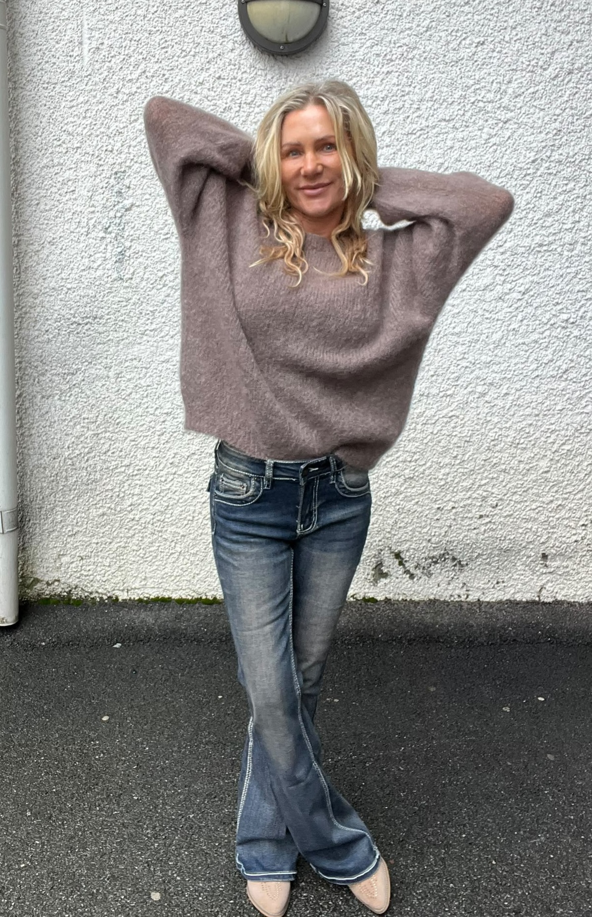 Kvinna med blont, lockigt hår står avslappnat med händerna bakom huvudet och ler mot kameran. Hon bär en mjuk stickad tröja, utsvängda jeans och ljusa skor, framför en vit putsad vägg utomhus.