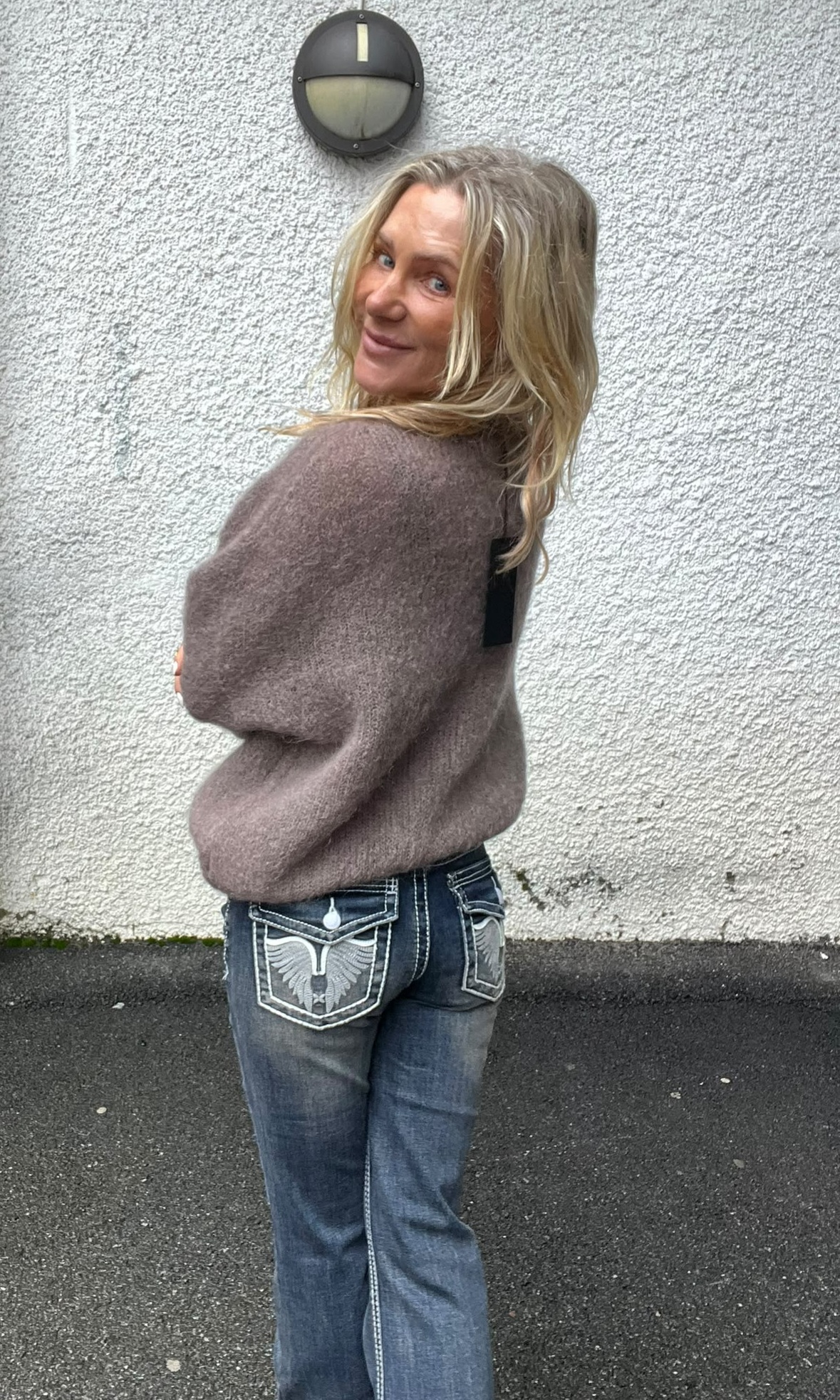 Blond kvinna står utomhus framför en vit, grovputsad vägg och vänder sig om med ett leende mot kameran. Hon bär en stickad brun tröja och blå jeans med broderade bakfickor; vägglampa ovanför.