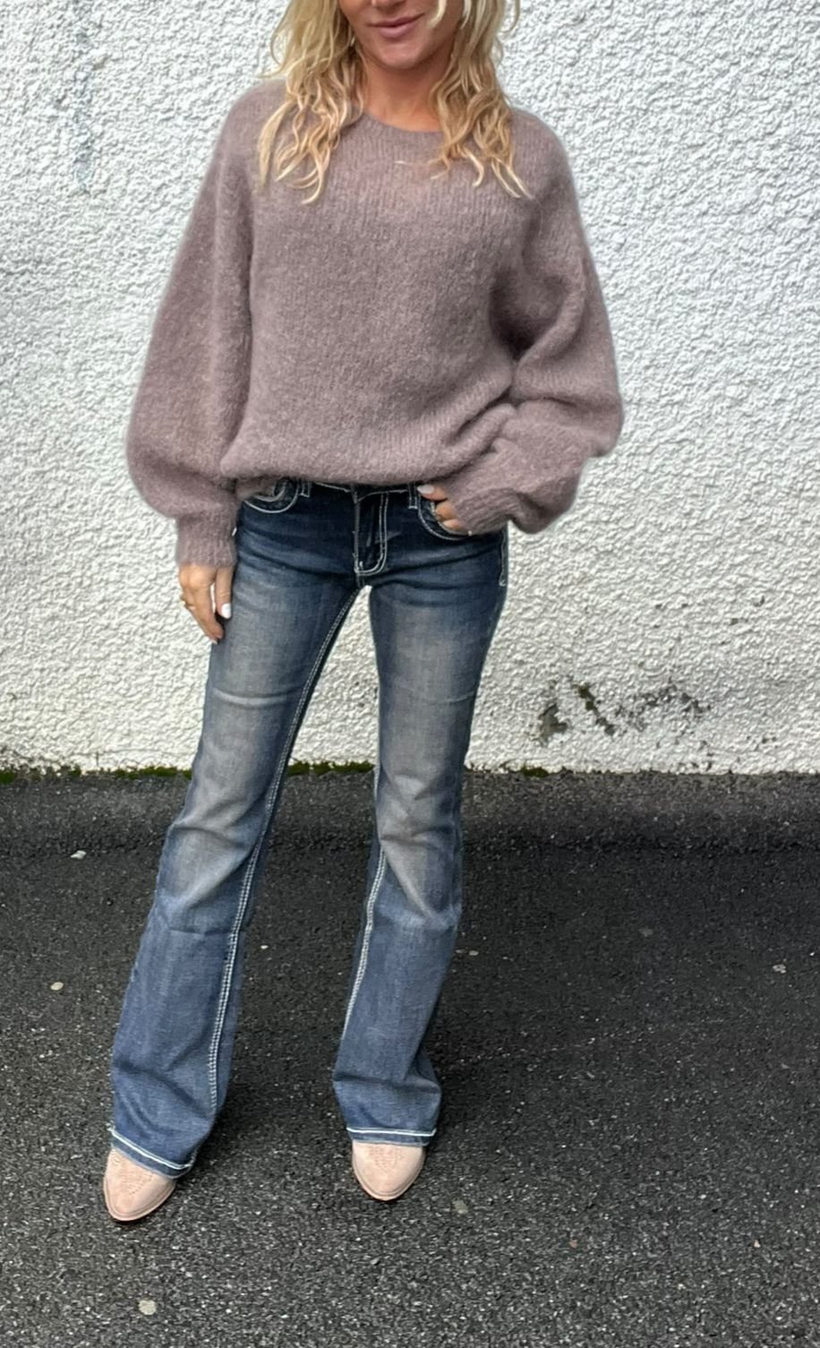 Kvinna står avslappnat mot en vit putsad vägg, med ena handen i jeansfickan. Hon bär en lila/grå stickad tröja med volymärmar, mörkblå utsvängda jeans och ljusa spetsiga skor på asfalt.