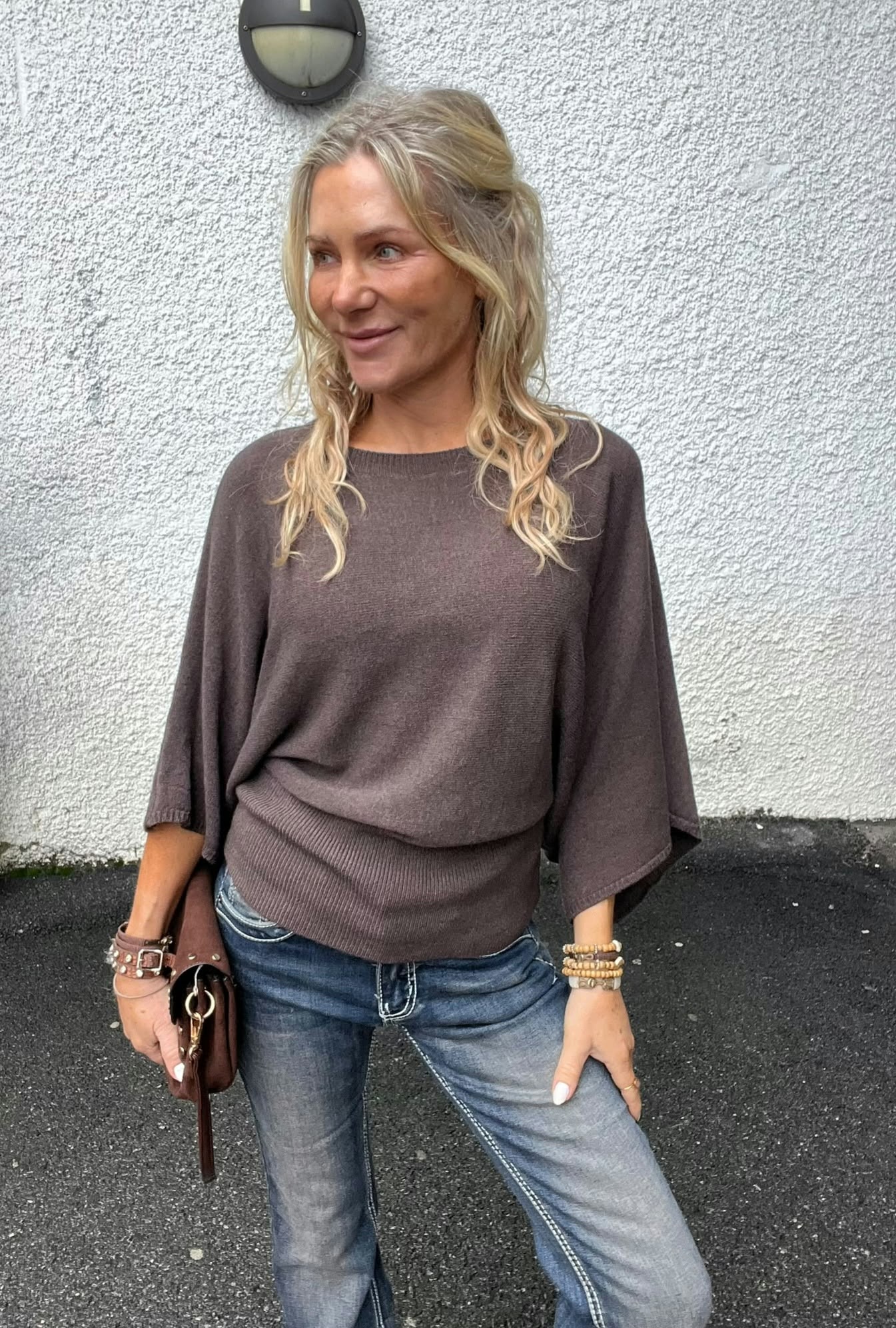 Kvinna med långt blont hår står lutad mot en vit, grovputsad vägg och tittar åt sidan med ett svagt leende. Hon bär brun stickad tröja och jeans, håller en brun väska; vägglampa syns ovanför.