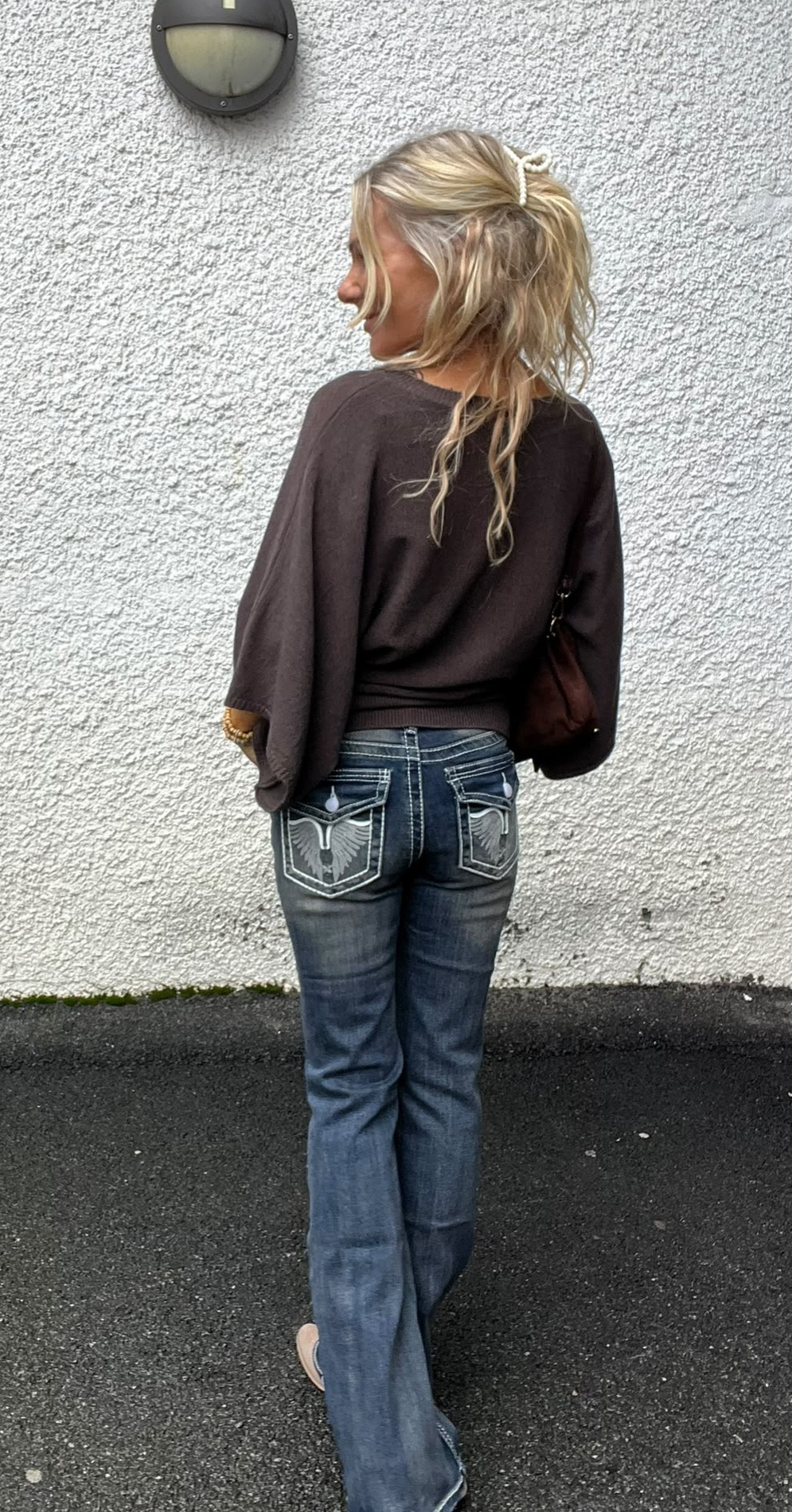 Person med blont, uppsatt hår står med ryggen mot kameran och tittar åt sidan, lutad mot en vit putsad vägg med rund vägglampa. Bär mörk tröja, utsvängda jeans med broderade bakfickor och liten axelväska.
