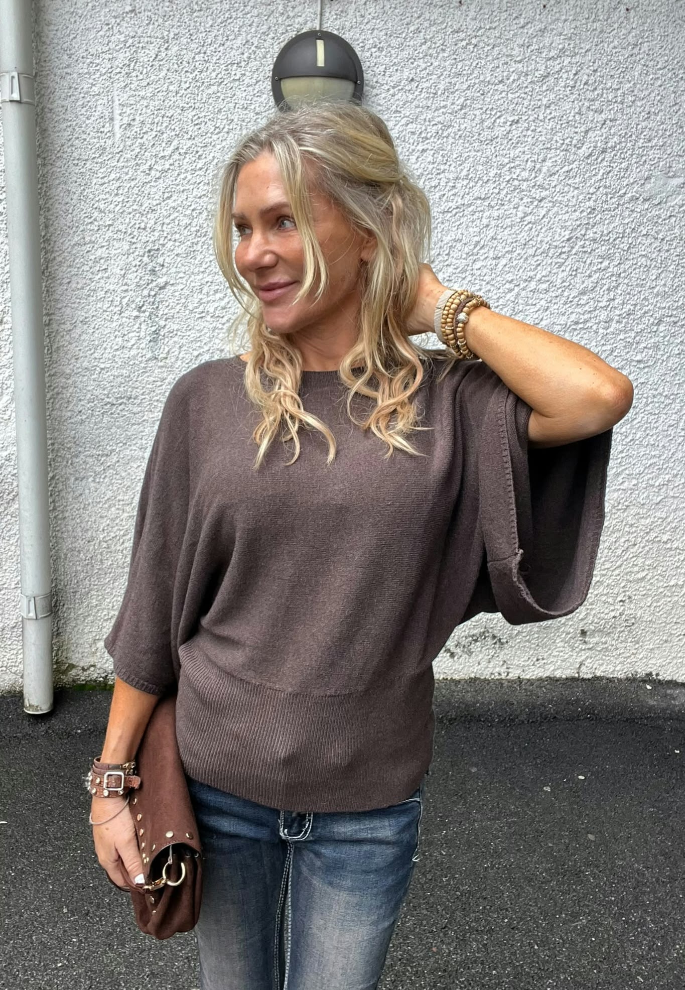 Blond kvinna står utomhus mot en vit, grovputsad vägg och tittar åt sidan med ett litet leende. Hon håller ena handen i nacken, bär brun stickad tröja, jeans, armband och en brun väska.
