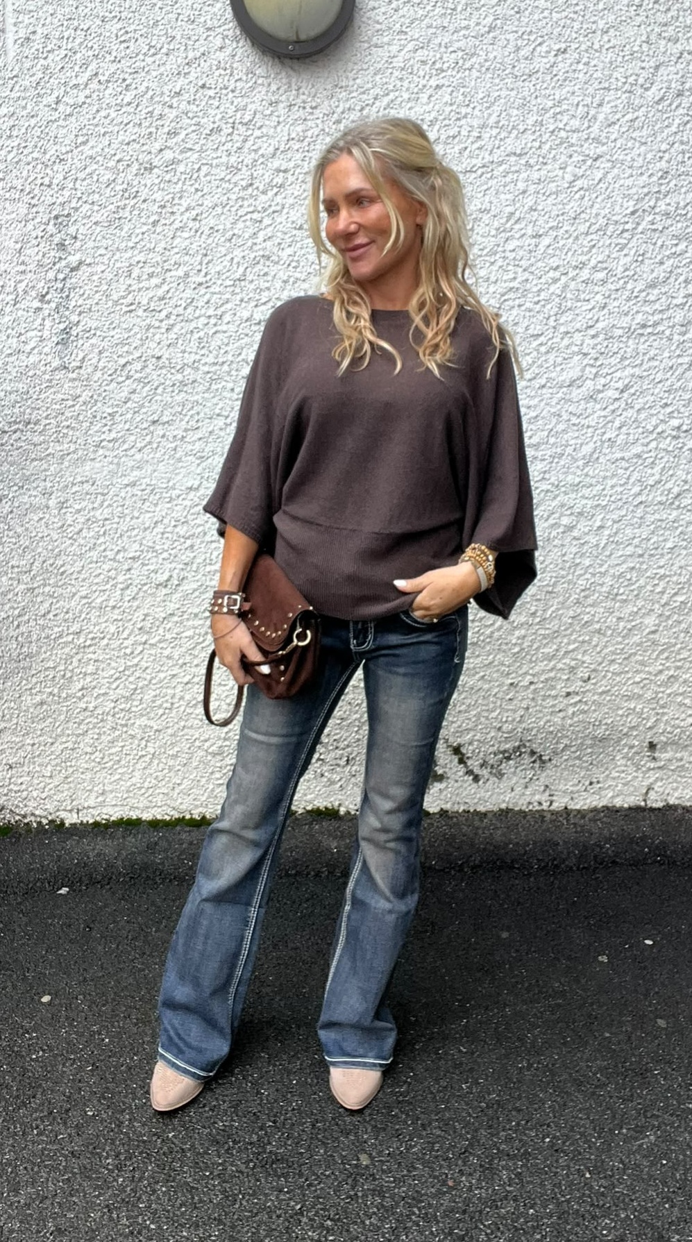Blond kvinna står avslappnat och tittar åt sidan, med ena handen i jeansfickan och en brun axelväska i den andra. Hon bär mörk ponchotröja, utsvängda jeans och ljusa skor mot vit putsad vägg.