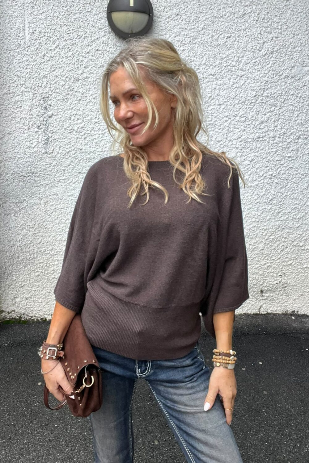 Blond kvinna står avslappnat mot en vit, grovputsad vägg och tittar åt sidan med ett svagt leende. Hon bär brun stickad tröja och jeans, håller en brun väska och har armband; vägglampa ovanför.
