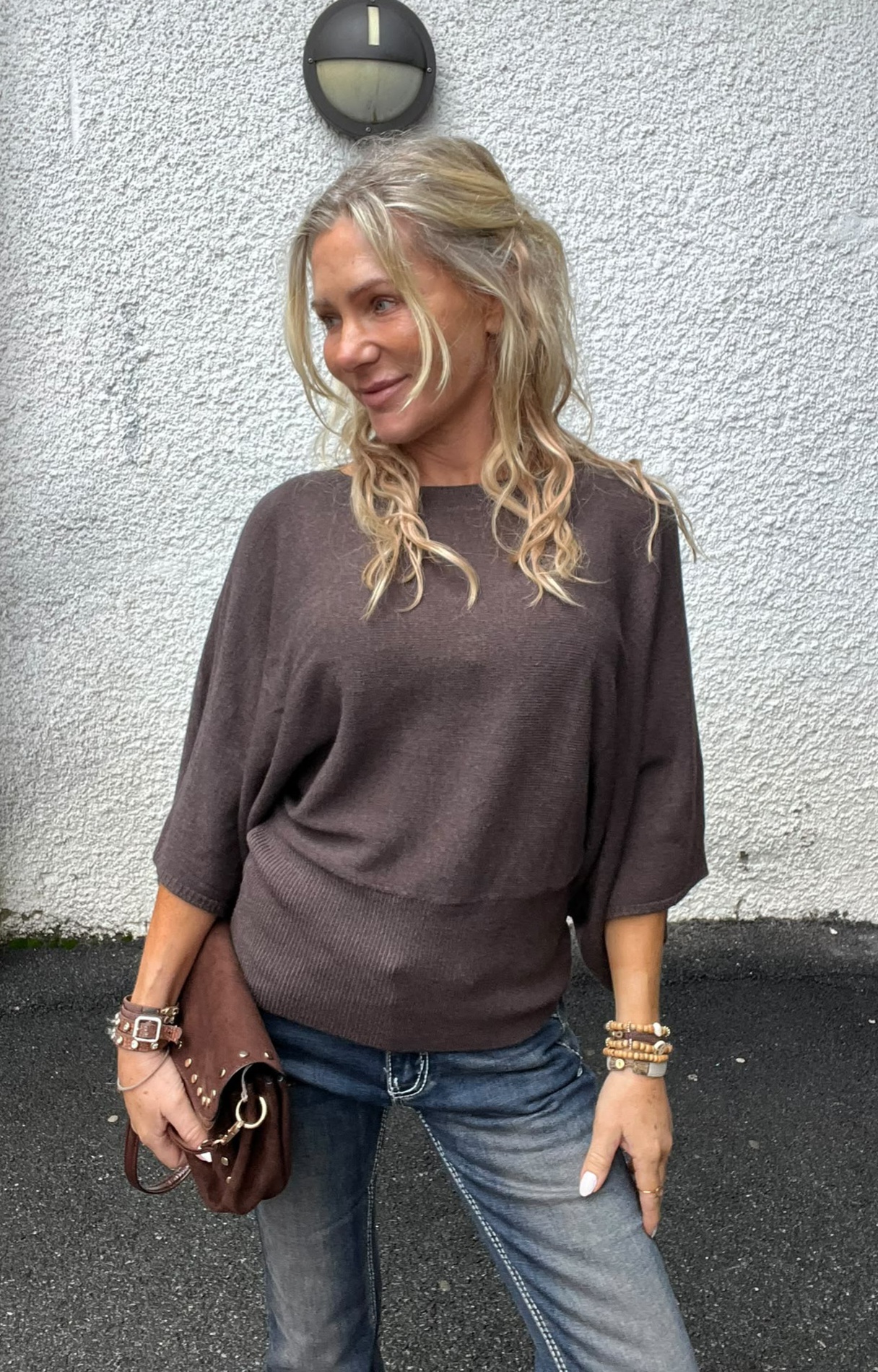 Blond kvinna står avslappnat mot en vit, grovputsad vägg och tittar åt sidan med ett svagt leende. Hon bär brun stickad tröja och jeans, håller en brun väska och har armband; vägglampa ovanför.