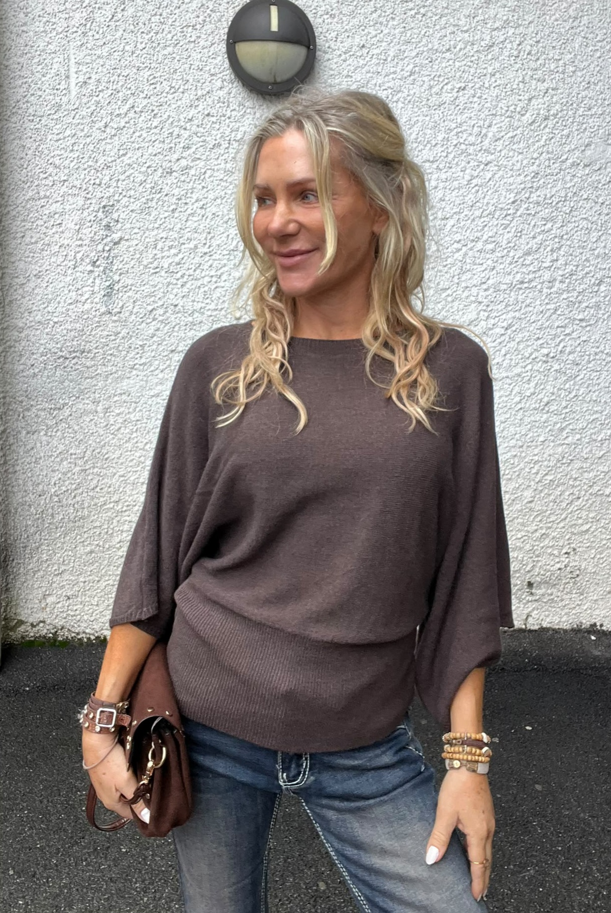 Blond kvinna står och ler medan hon tittar åt sidan, klädd i brun stickad tröja med vida ärmar och jeans. Hon håller en brun väska och bär armband, framför en vit putsad vägg med rund vägglampa.