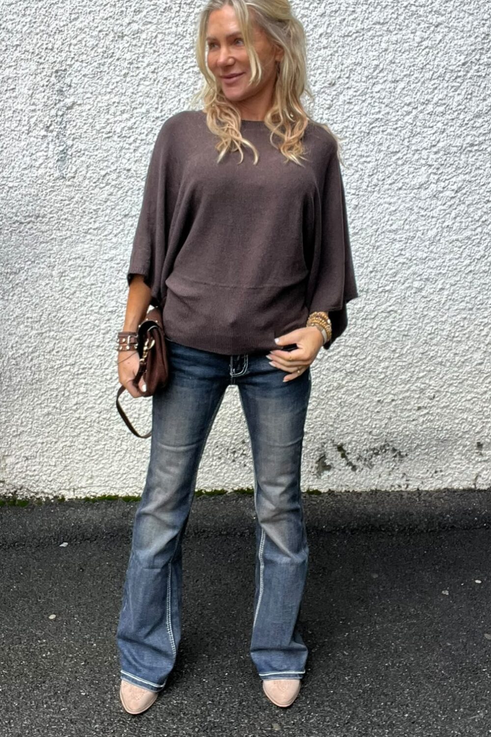 Blond kvinna står avslappnat mot en vit, grovputsad vägg och ler åt sidan. Hon håller en liten brun handväska och bär mörkbrun, vid tröja samt blå bootcut-jeans och ljusa skor på asfalt.