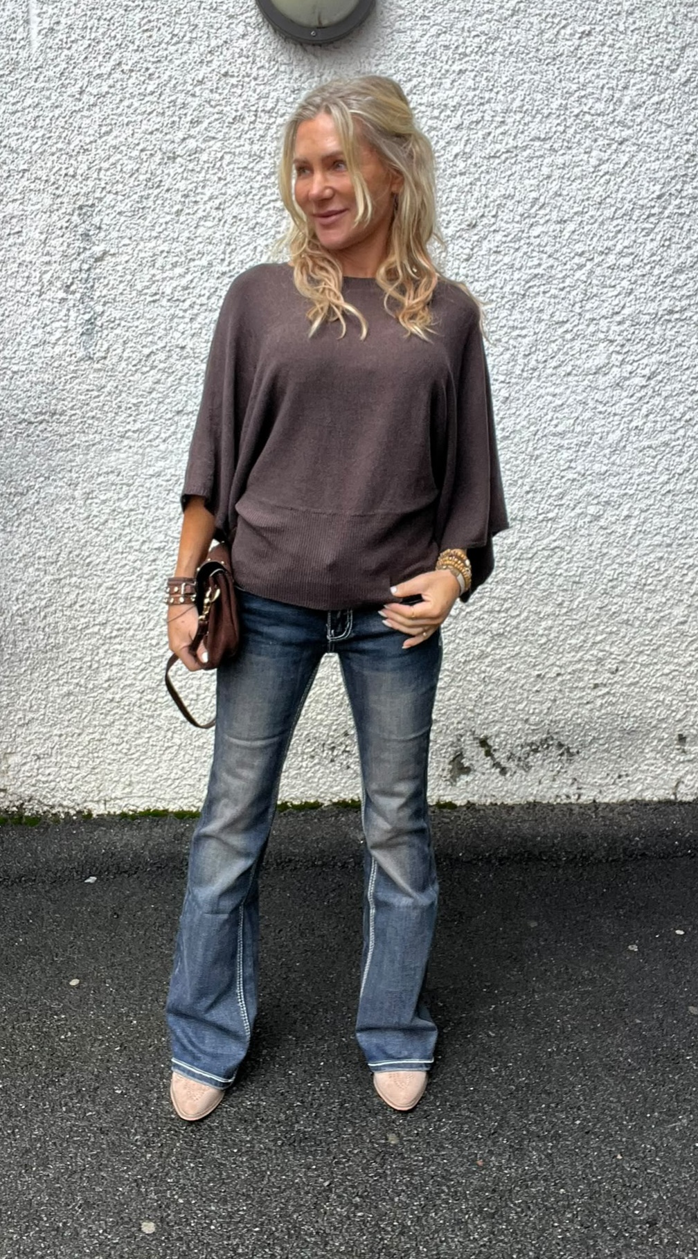 Blond kvinna står avslappnat mot en vit, grovputsad vägg och ler åt sidan. Hon håller en liten brun handväska och bär mörkbrun, vid tröja samt blå bootcut-jeans och ljusa skor på asfalt.