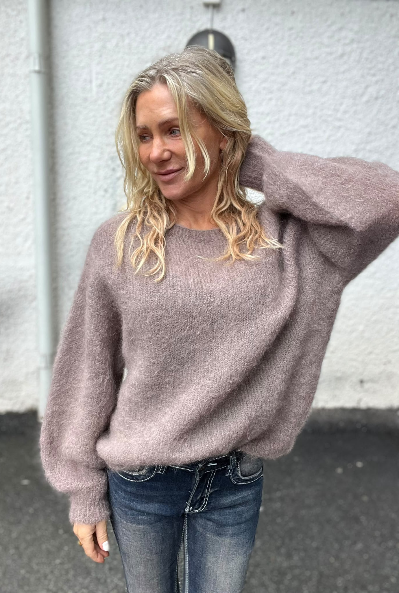 Kvinna med långt, blont vågigt hår står avslappnat och ler svagt, med ena handen bakom nacken. Hon bär en lila stickad tröja och jeans, fotograferad utomhus mot en ljus vägg.
