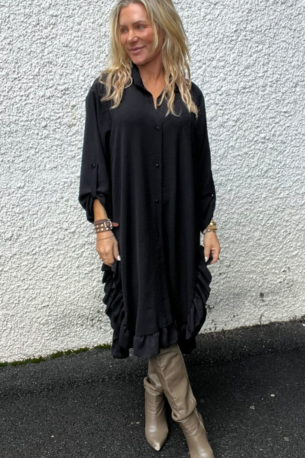 Kvinna med långt blont hår står avslappnat mot en vit, grovputsad vägg och tittar åt sidan. Hon bär en svart, lös tunikaklänning med volangkant, smycken på handlederna och beige, spetsiga stövlar på asfalt.