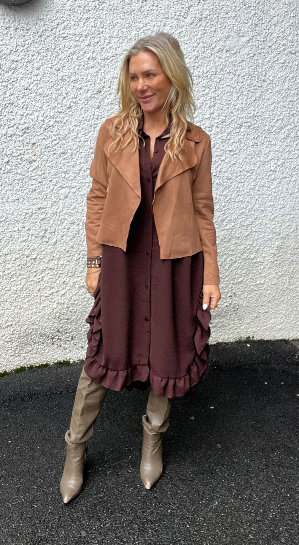 Blond kvinna står lutad mot en vit, grov putsad vägg och ler åt sidan. Hon bär vinröd klänning med knappar och volangkant, camel-färgad kort jacka samt beige spetsiga stövlar på mörk asfalt.