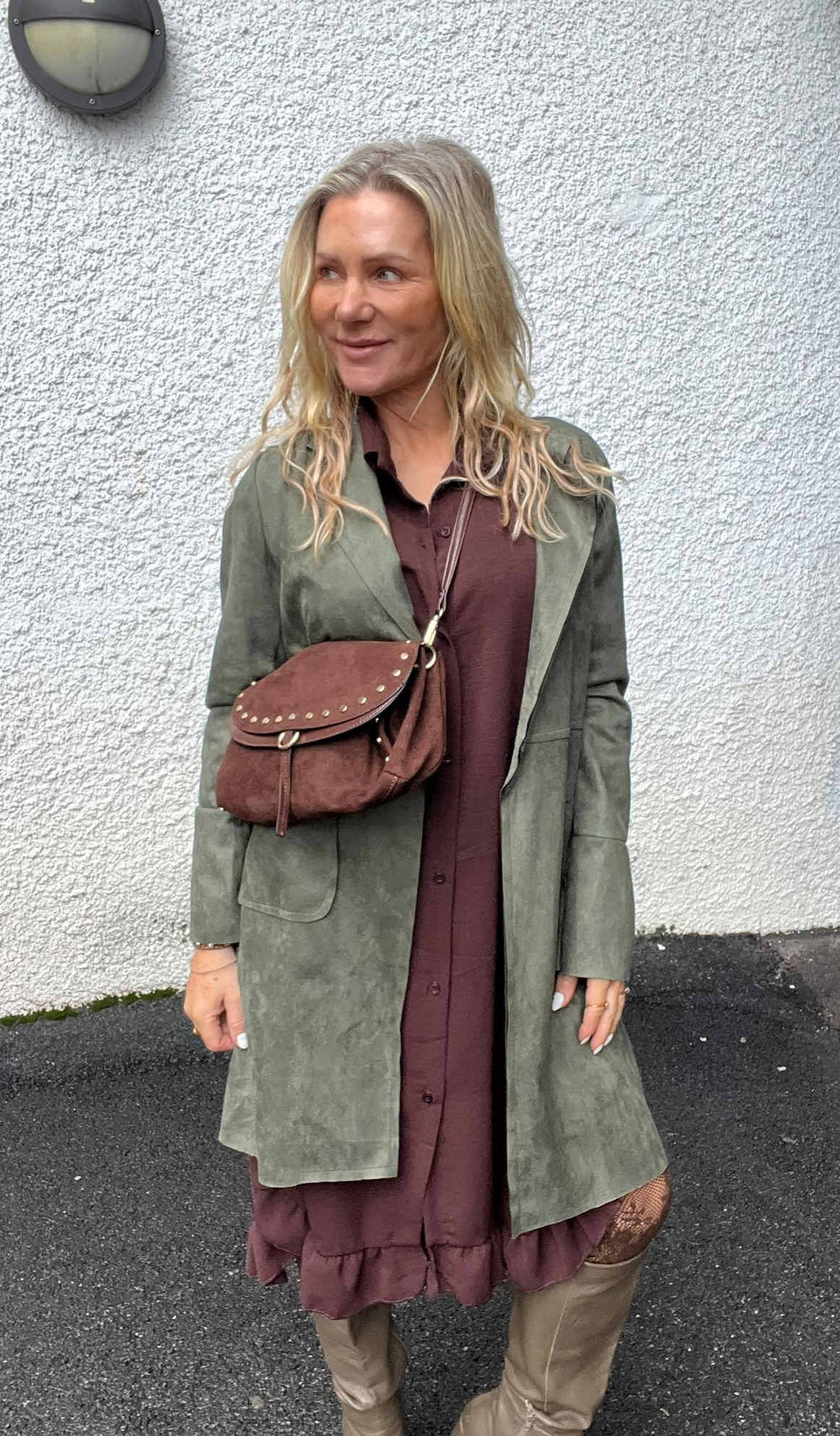 Blond kvinna står utomhus mot en vit, grovputsad vägg och tittar åt sidan med ett litet leende. Hon bär olivgrön kappa över vinröd klänning, brun axelväska med nitar samt beige höga stövlar.