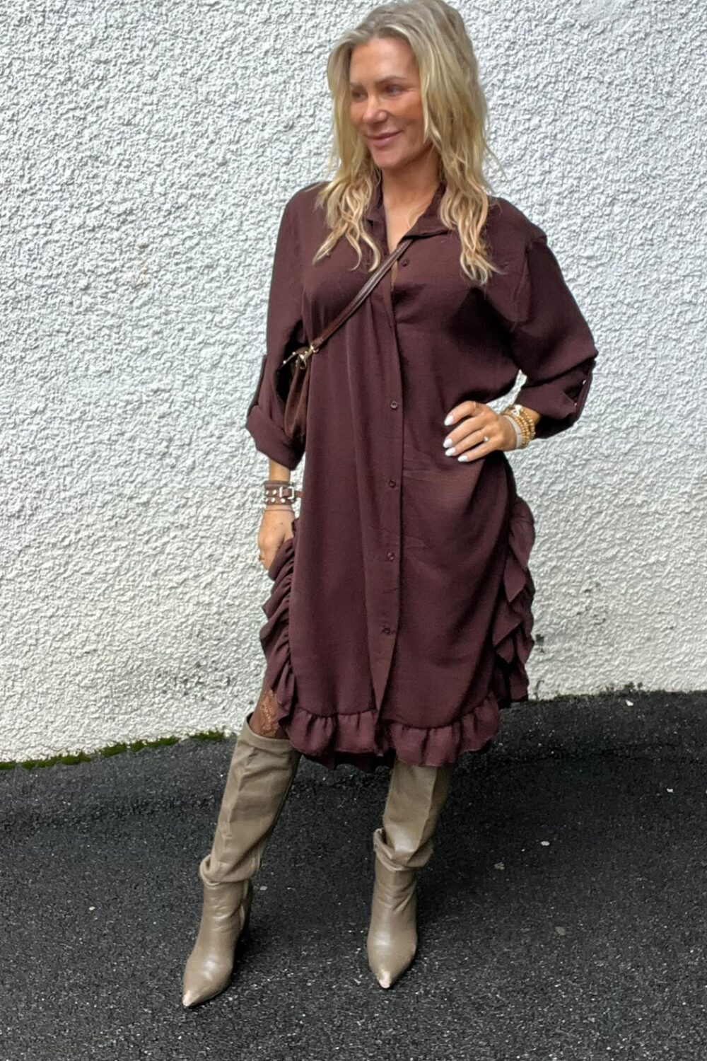 Blond kvinna poserar med handen på höften och ler, klädd i vinröd skjortklänning med volanger och knappar. Hon bär beige knähöga stövlar och brun axelväska, och står utomhus mot en vit, grov vägg.