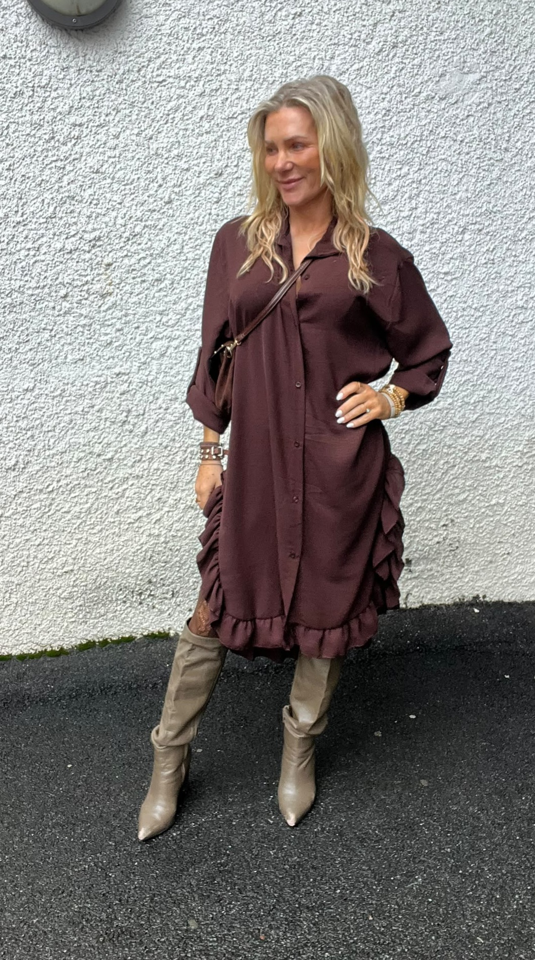 Blond kvinna poserar med handen på höften och ler, klädd i vinröd skjortklänning med volanger och knappar. Hon bär beige knähöga stövlar och brun axelväska, och står utomhus mot en vit, grov vägg.