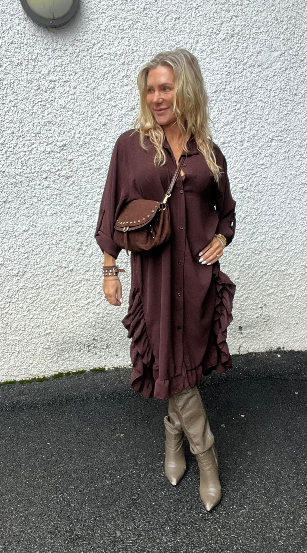 Blond kvinna står och ler vid en vit, grovputsad vägg, vänd åt sidan. Hon bär en mörkbrun skjortklänning med volanger, brun axelväska med nitar och beige knähöga stövlar, på asfalterad mark utomhus.