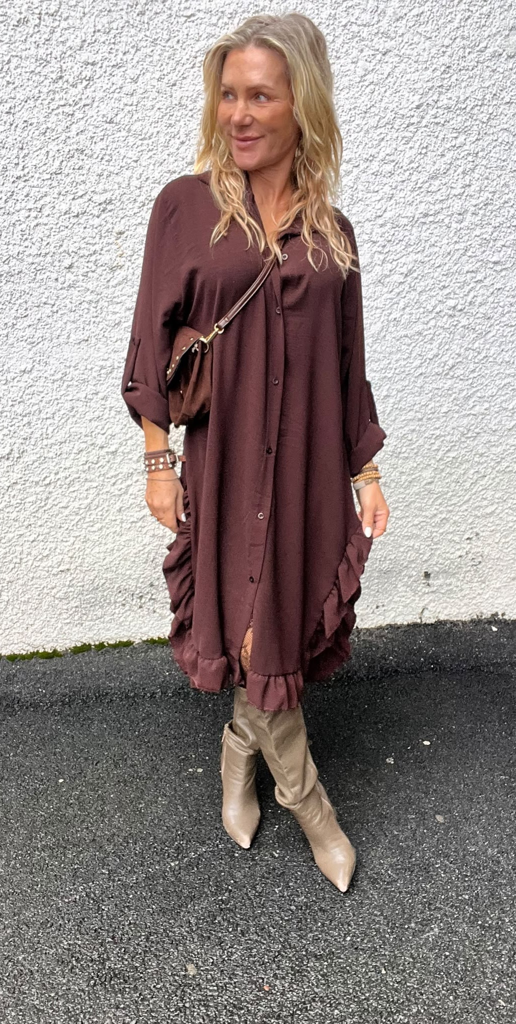 Blond kvinna står utomhus mot en vit, grovputsad vägg och ler medan hon tittar åt sidan. Hon bär en lång vinröd skjortklänning med volangkant, axelväska och beige knähöga stövlar.